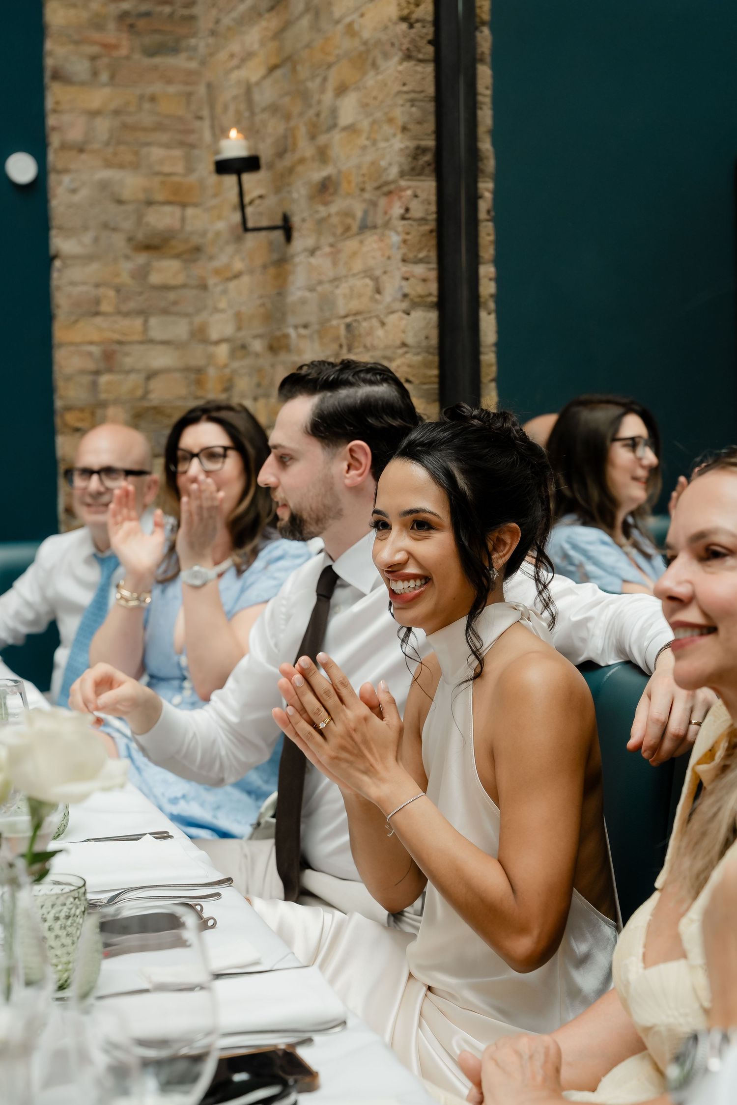 Smiling bride in white dress clapping at wedding reception with guests.