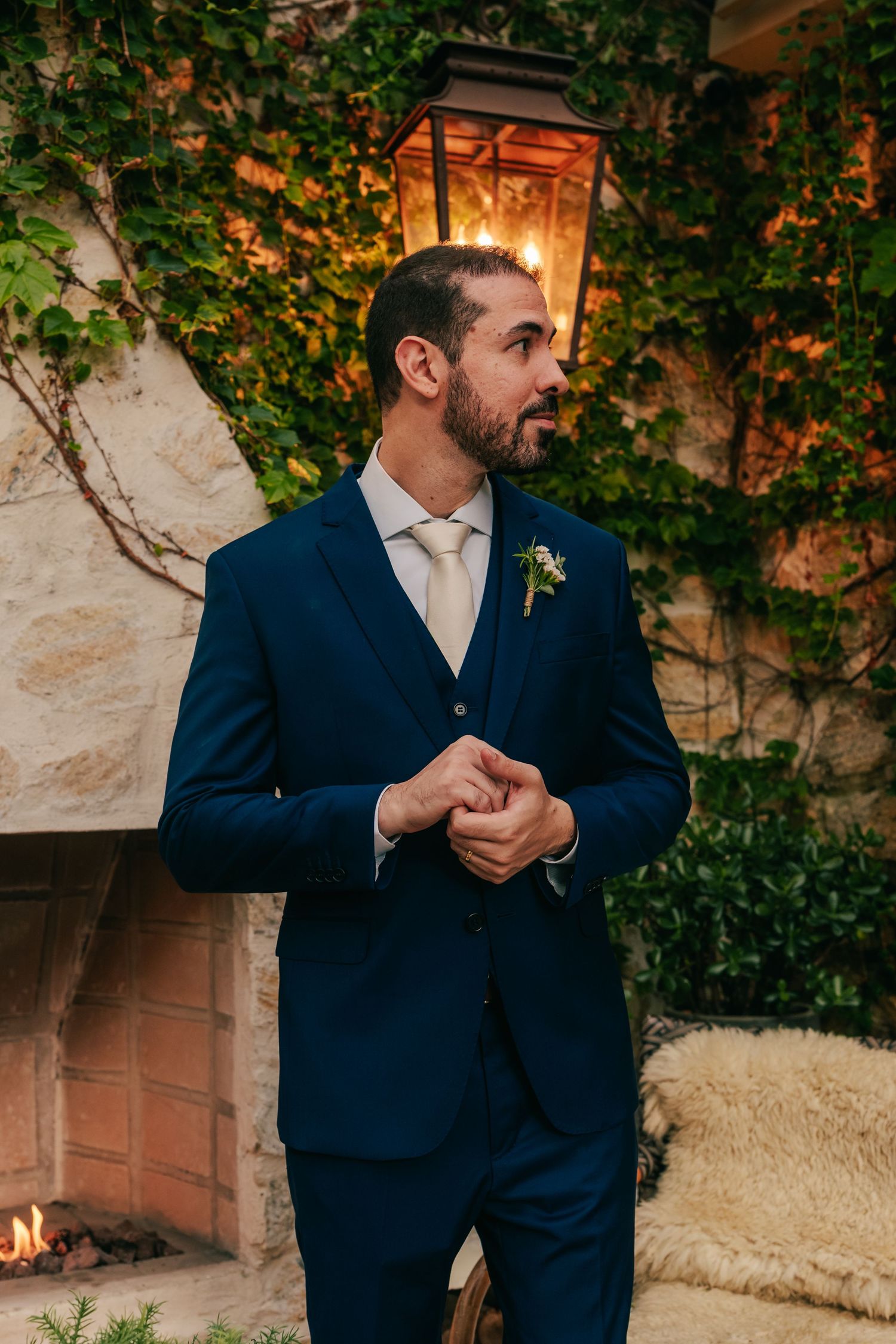 Dapper groom in navy blue three-piece suit with boutonniere stands by stone fireplace under warm lantern light.