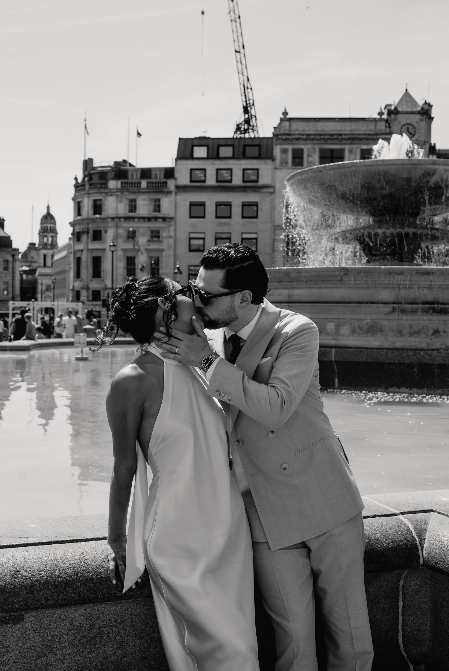 Couple sharing a romantic kiss in front of Trafalgar Square fountain on their wedding day in London.
