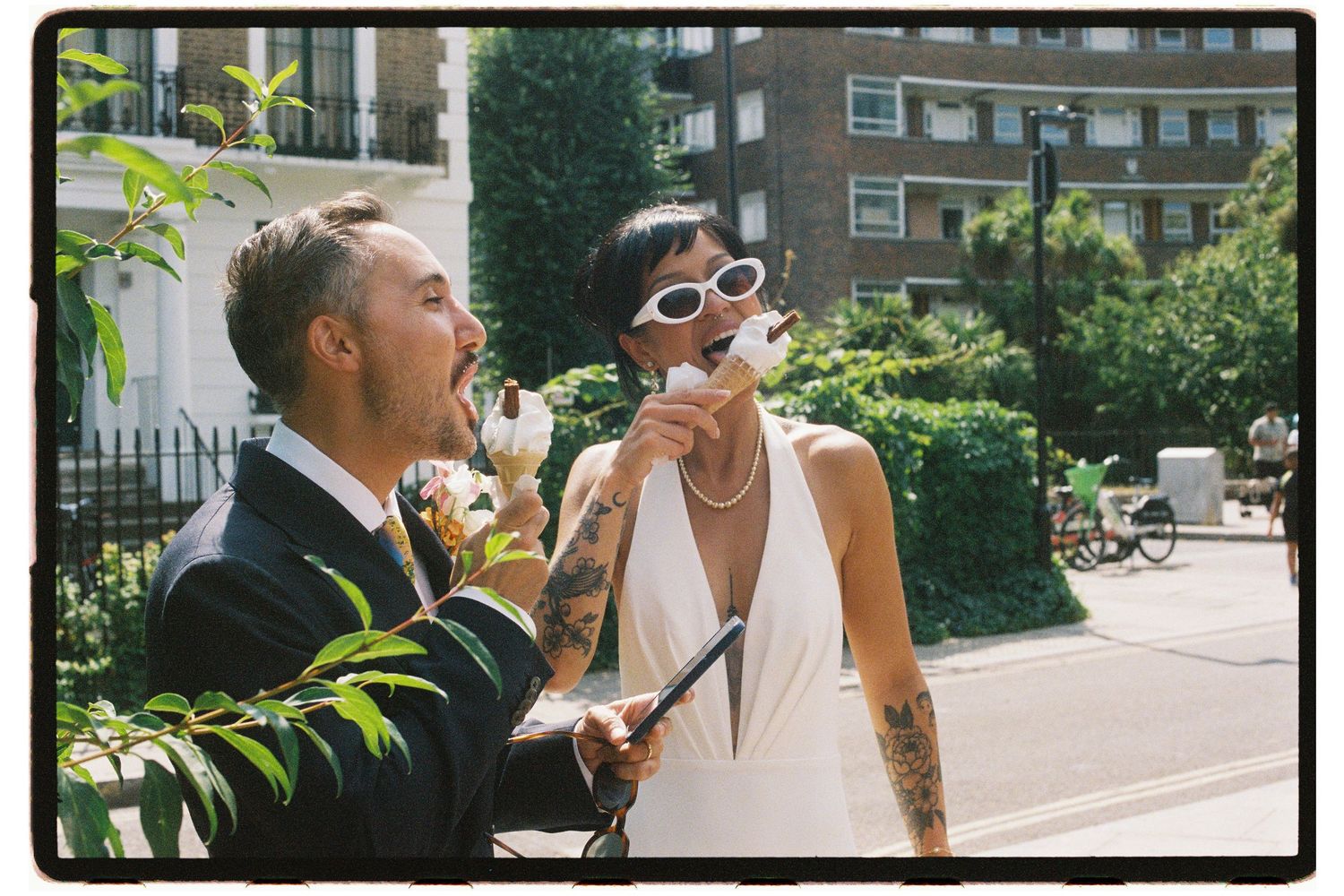Tattooed bride and groom enjoy ice cream cones together on a sunny city street.