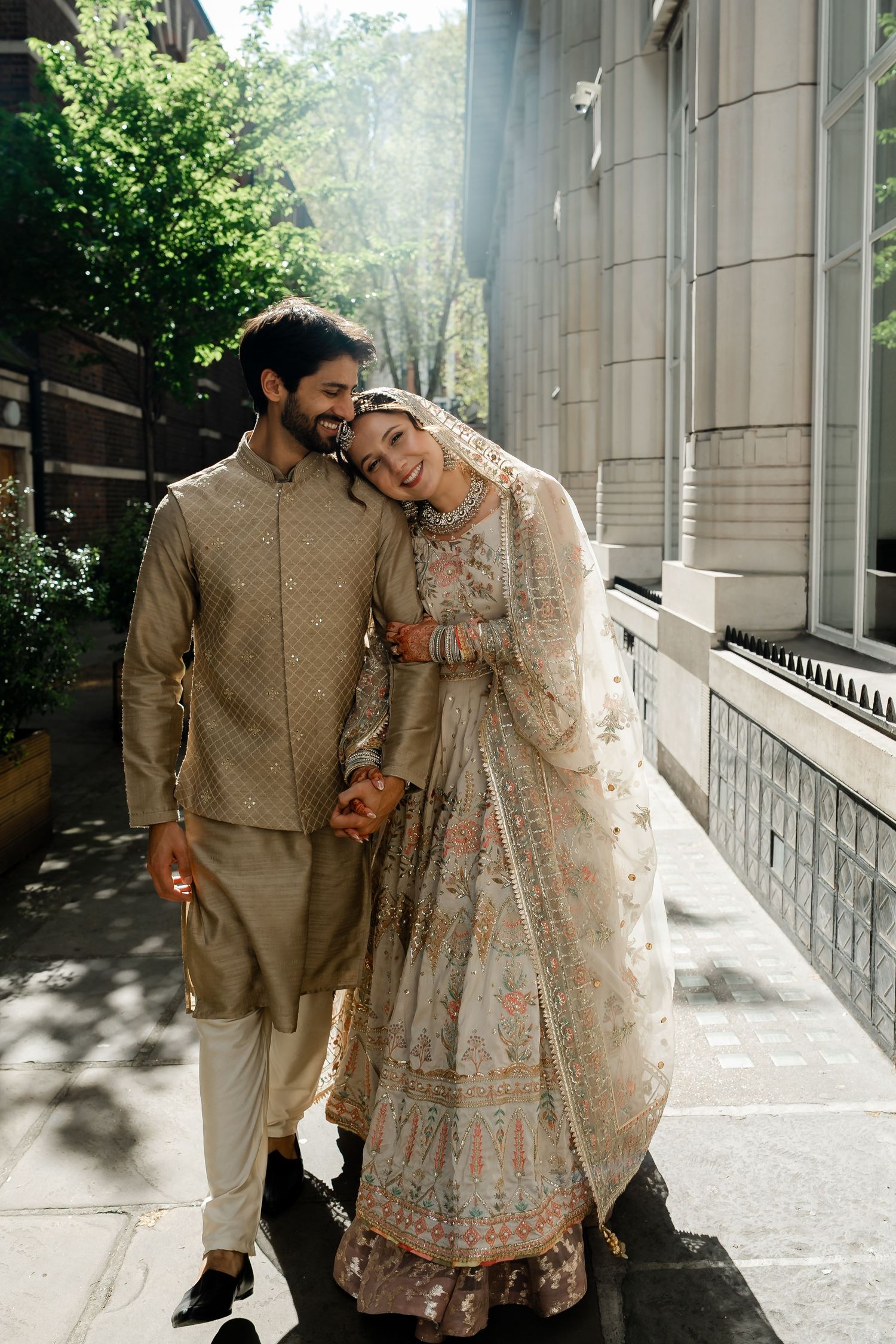 South Asian couple in traditional wedding attire walking hand in hand on sunny city street smiling joyfully.