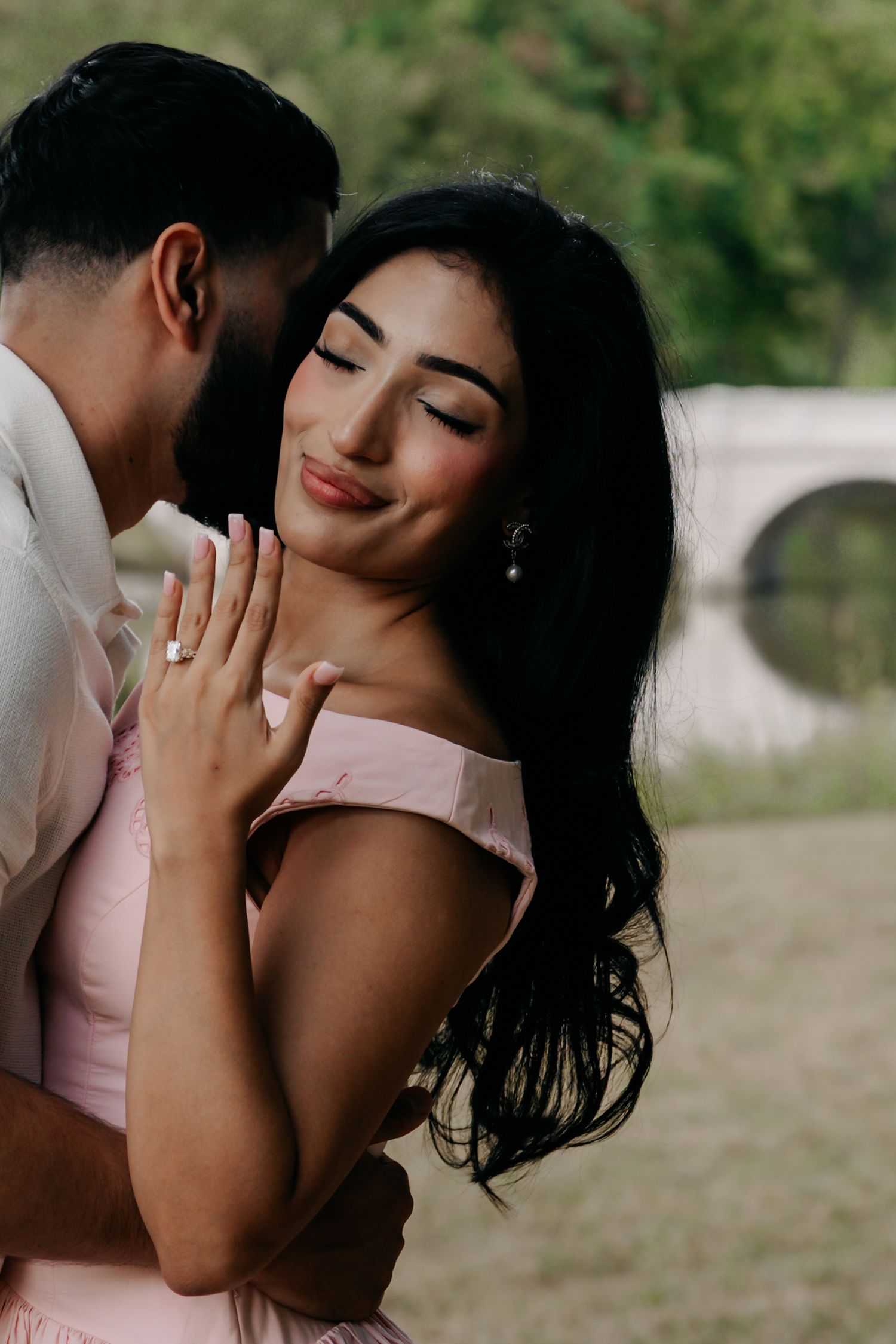 Happy woman smiles as her partner whispers in her ear, proudly displaying her diamond engagement ring outdoors.