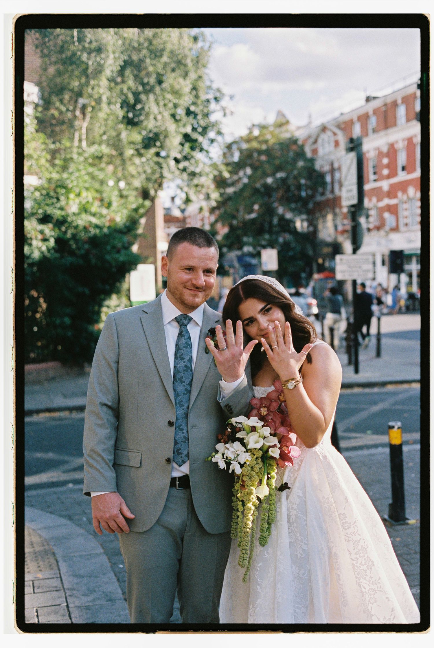 Newlyweds showing wedding rings on a sunny street, bride holding bouquet, groom in gray suit, urban background.