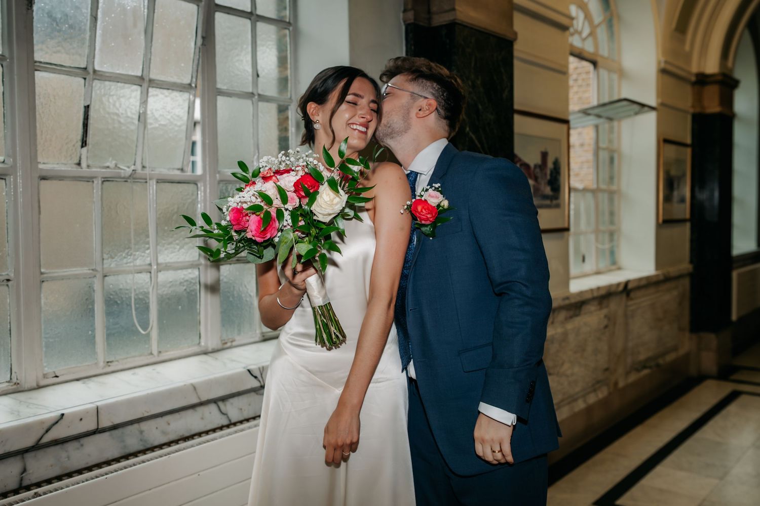 Bride holding pink bouquet smiles as groom in navy suit kisses her cheek in elegant indoor wedding venue.