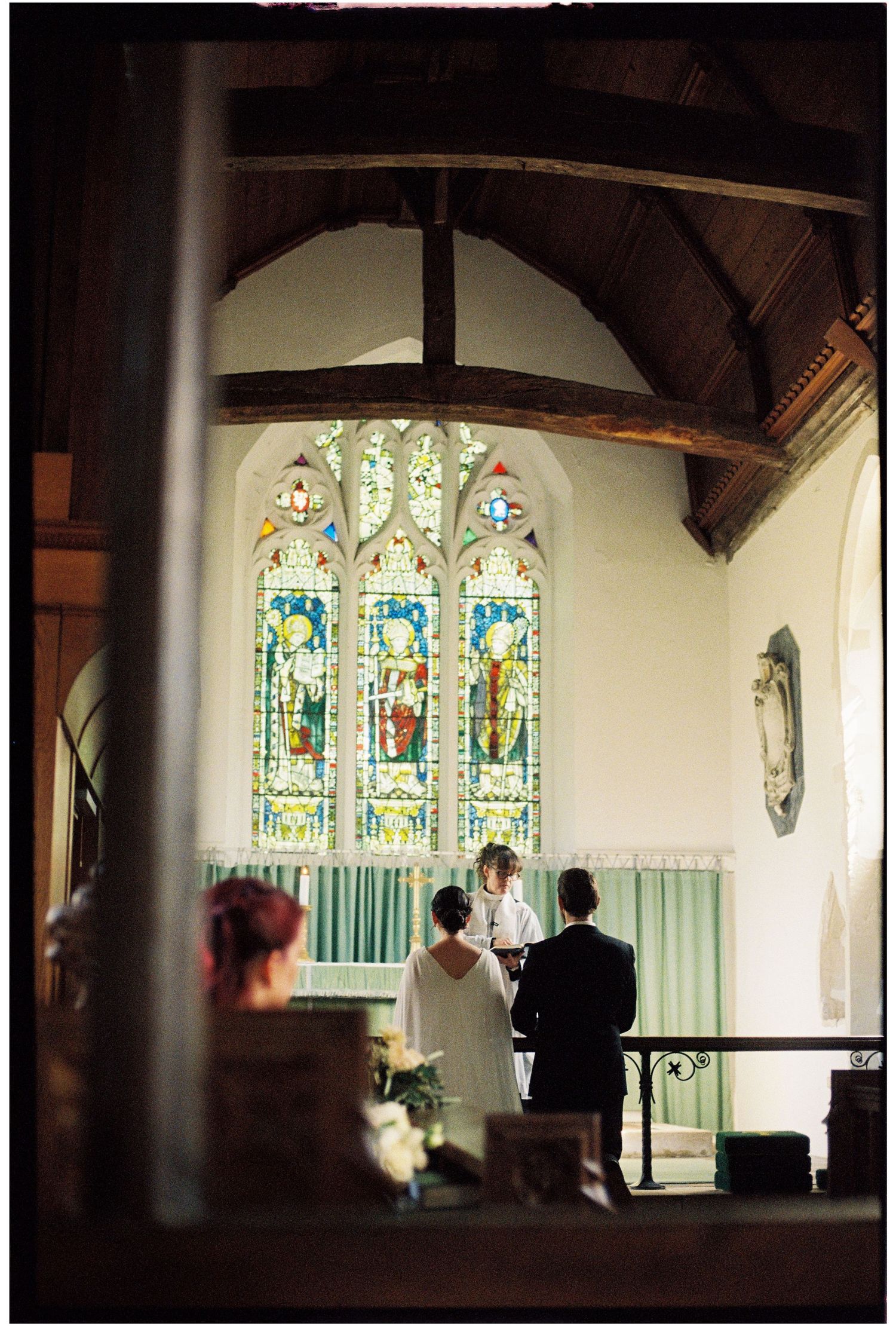 Couple exchanging vows at church altar with colorful stained glass windows and wooden beam ceiling.