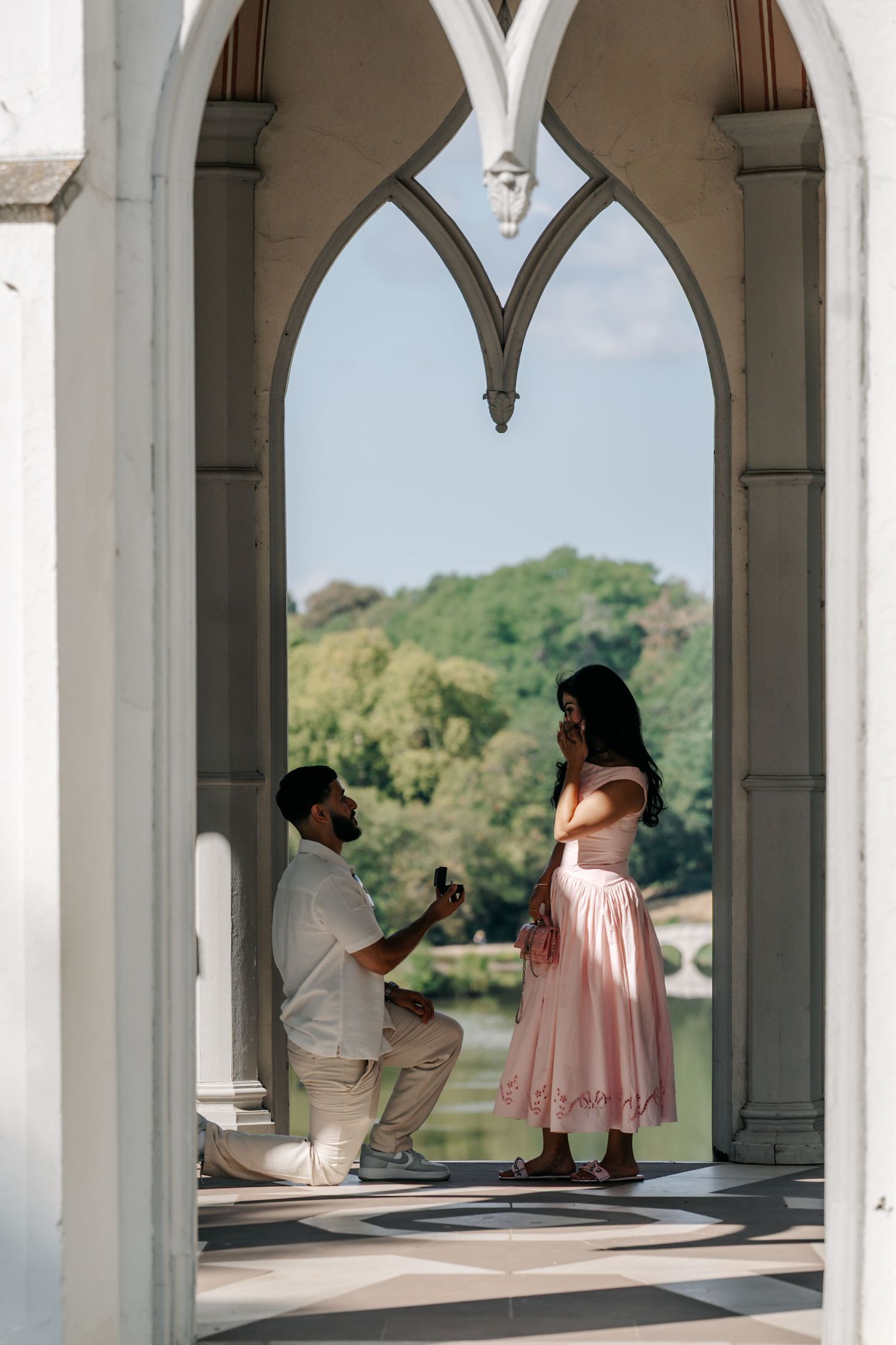 Man proposes on one knee to woman in pink dress under Gothic arch with lake backdrop.