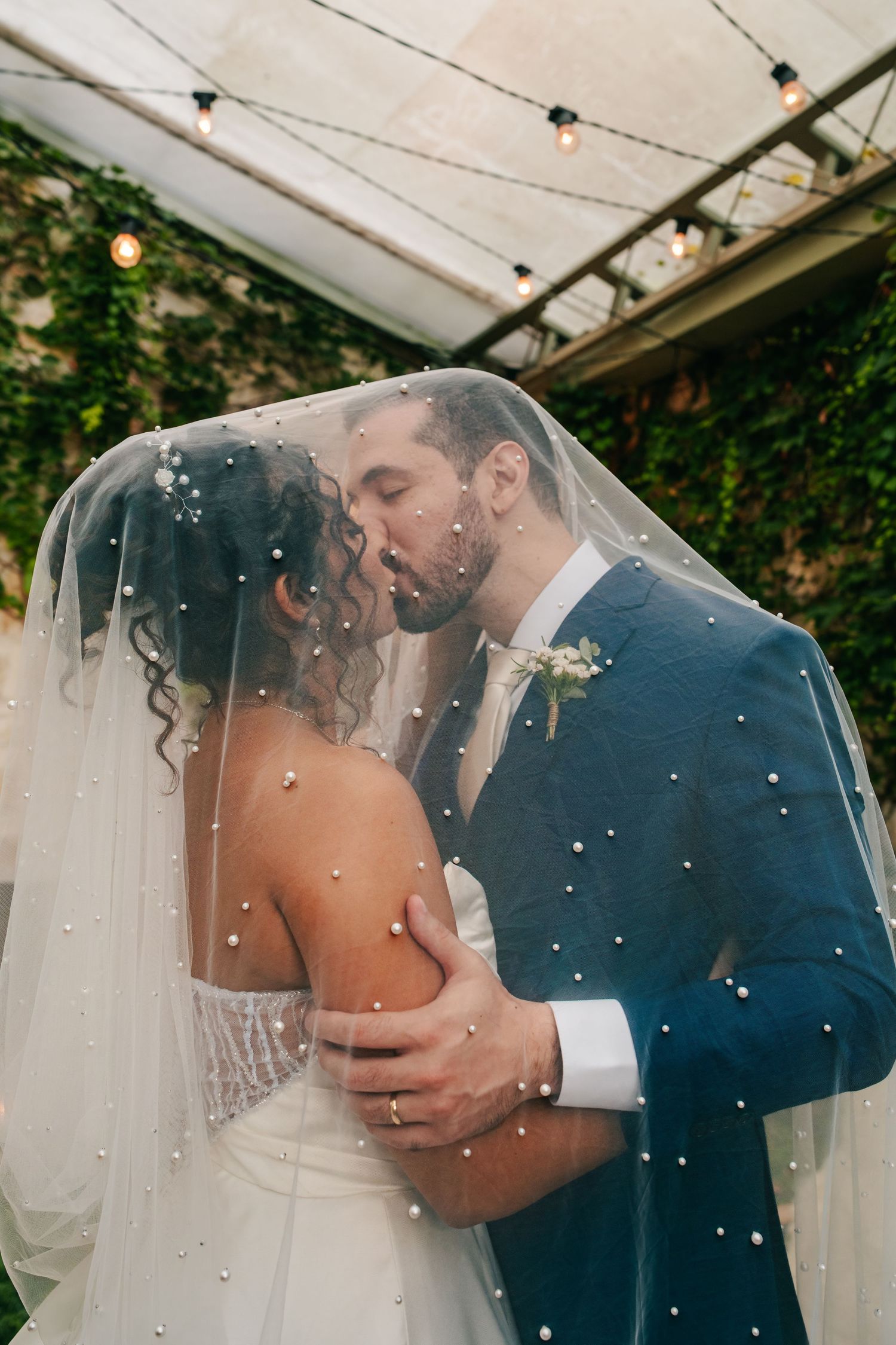 Bride and groom share a romantic kiss under pearl-adorned wedding veil outdoors.