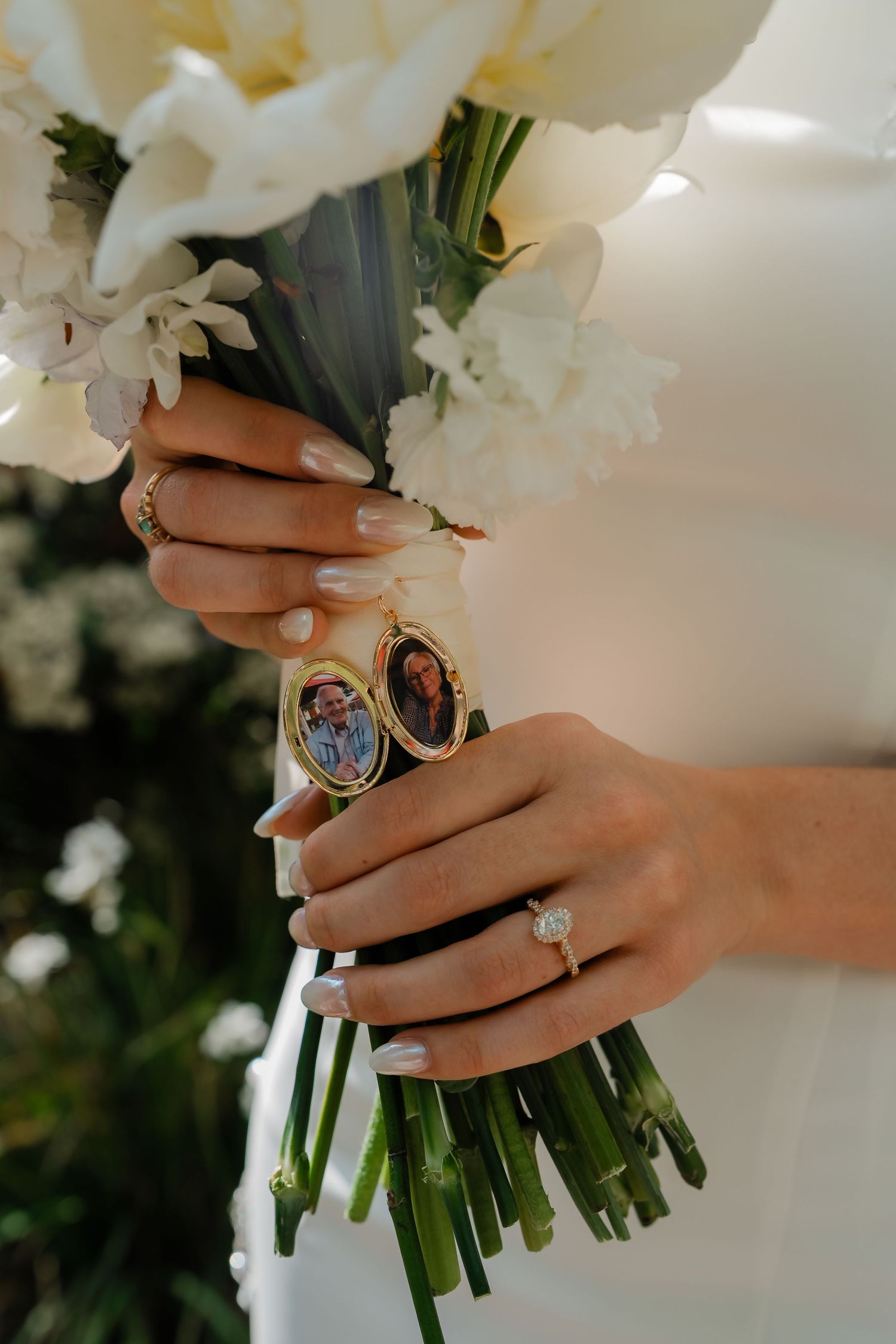 Bride holds white floral bouquet adorned with gold locket charms and diamond engagement ring.