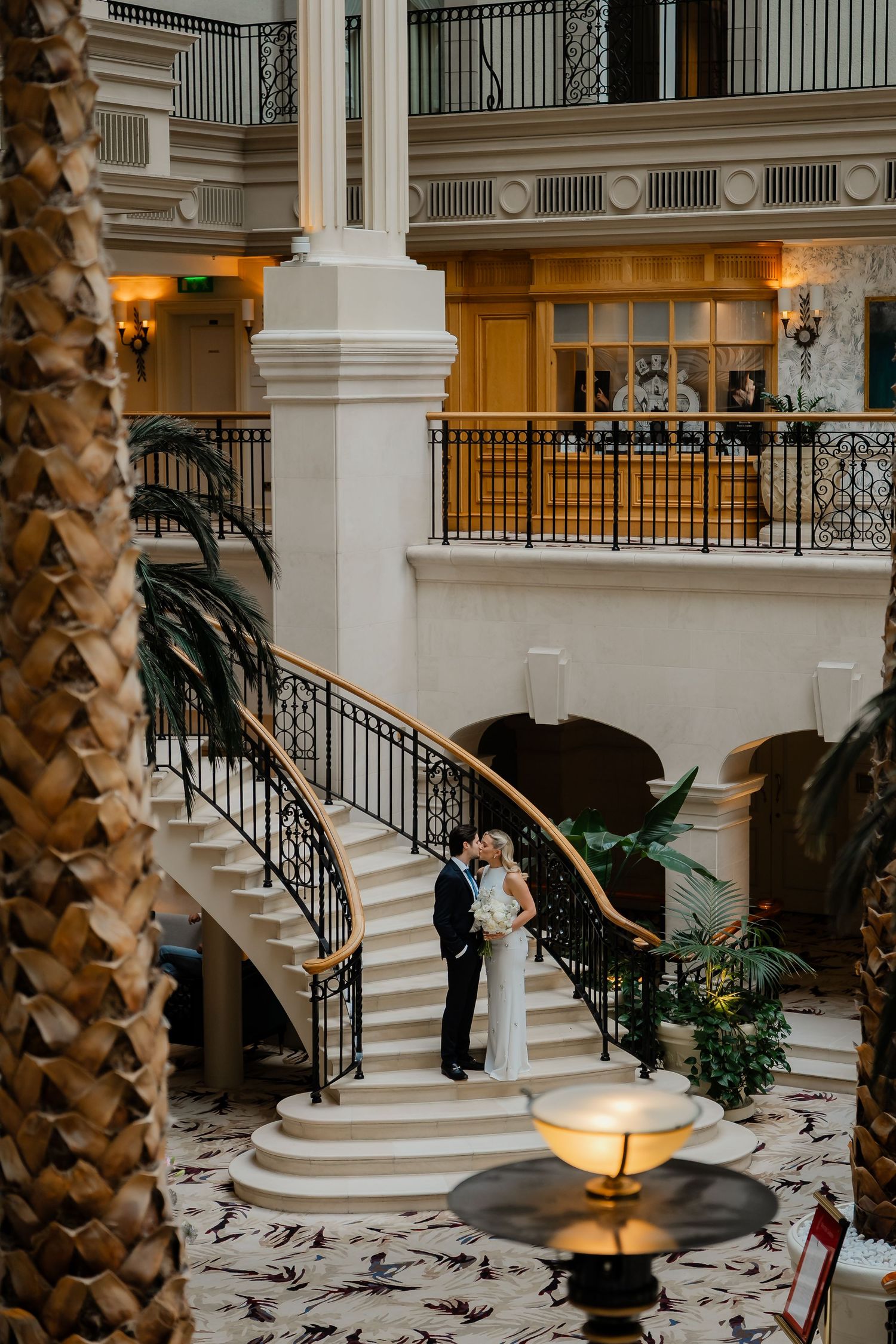 Bride and groom kissing on a sweeping grand staircase in a luxurious hotel atrium with palm trees.