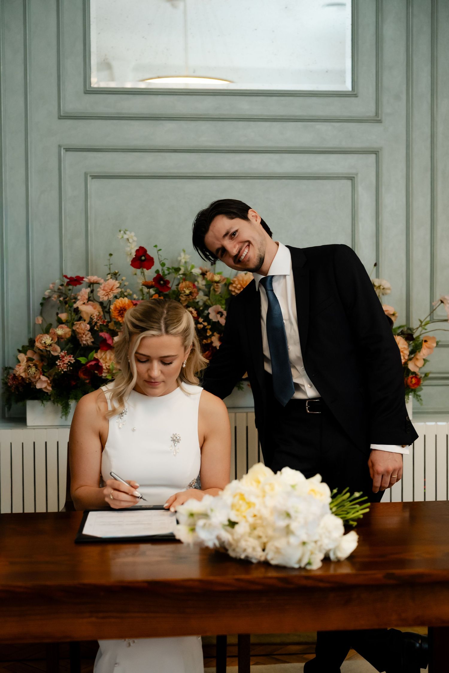 Bride signs marriage register as smiling groom leans in beside floral arrangement.