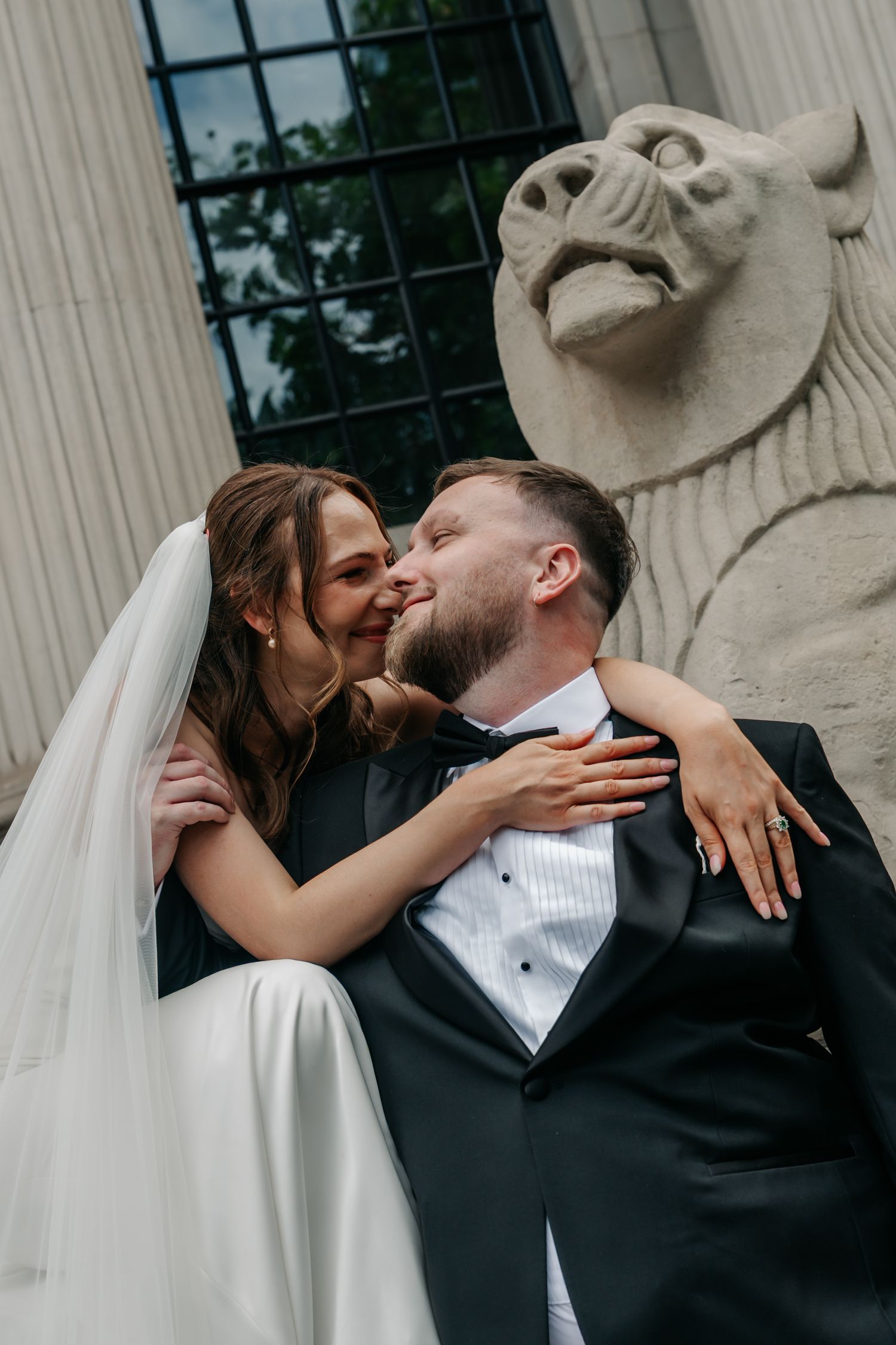 Bride and groom share an intimate moment beside a stone lion sculpture at a classic building.