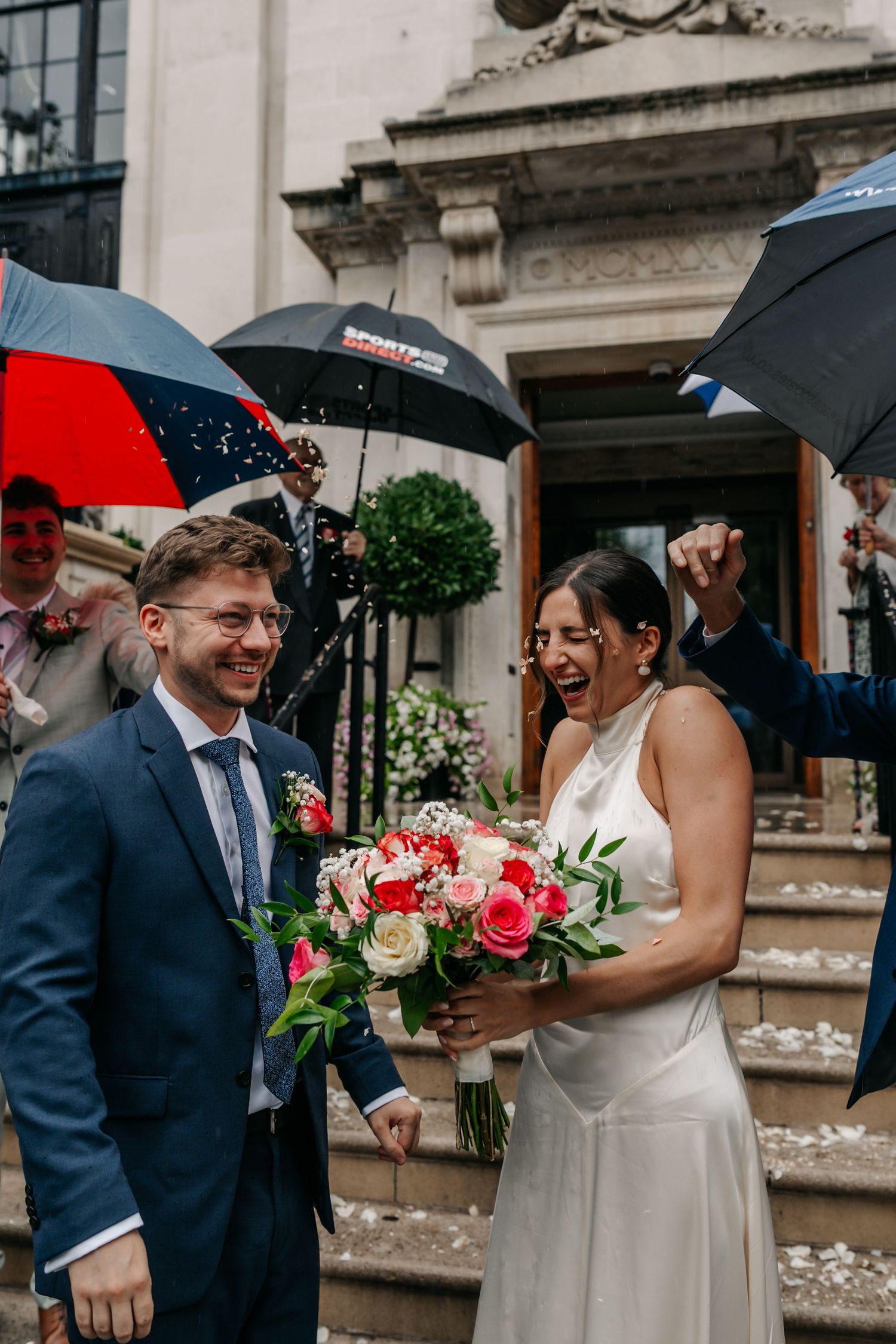 Joyful newlyweds exit wedding venue in rain, bride holding pink bouquet, guests with umbrellas throwing confetti.
