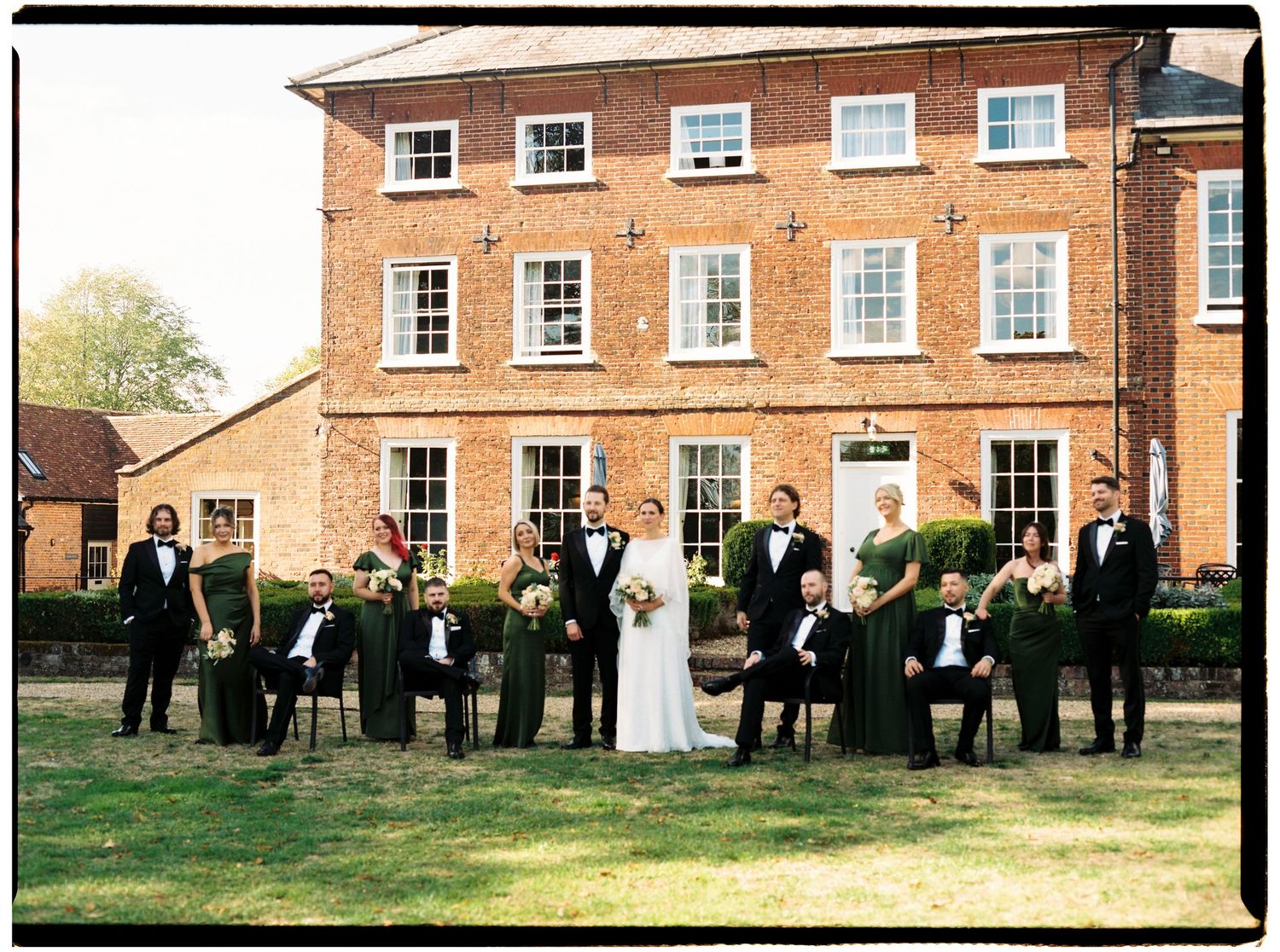 Wedding party posing in front of a red brick Georgian venue, bridesmaids in emerald green gowns, groomsmen in black tuxedos.