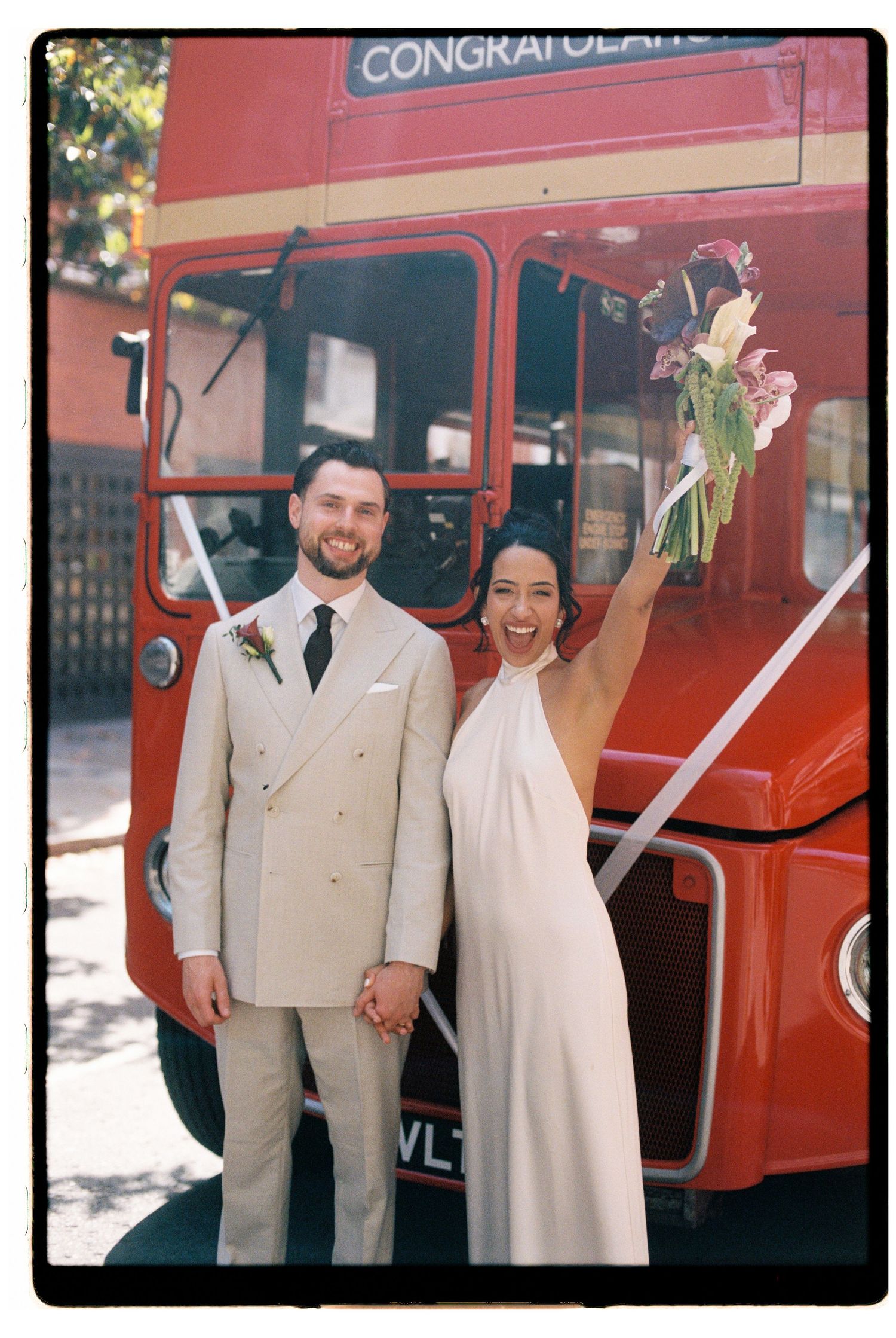 Smiling newlyweds posing before iconic red London double-decker bus with bride raising bouquet.