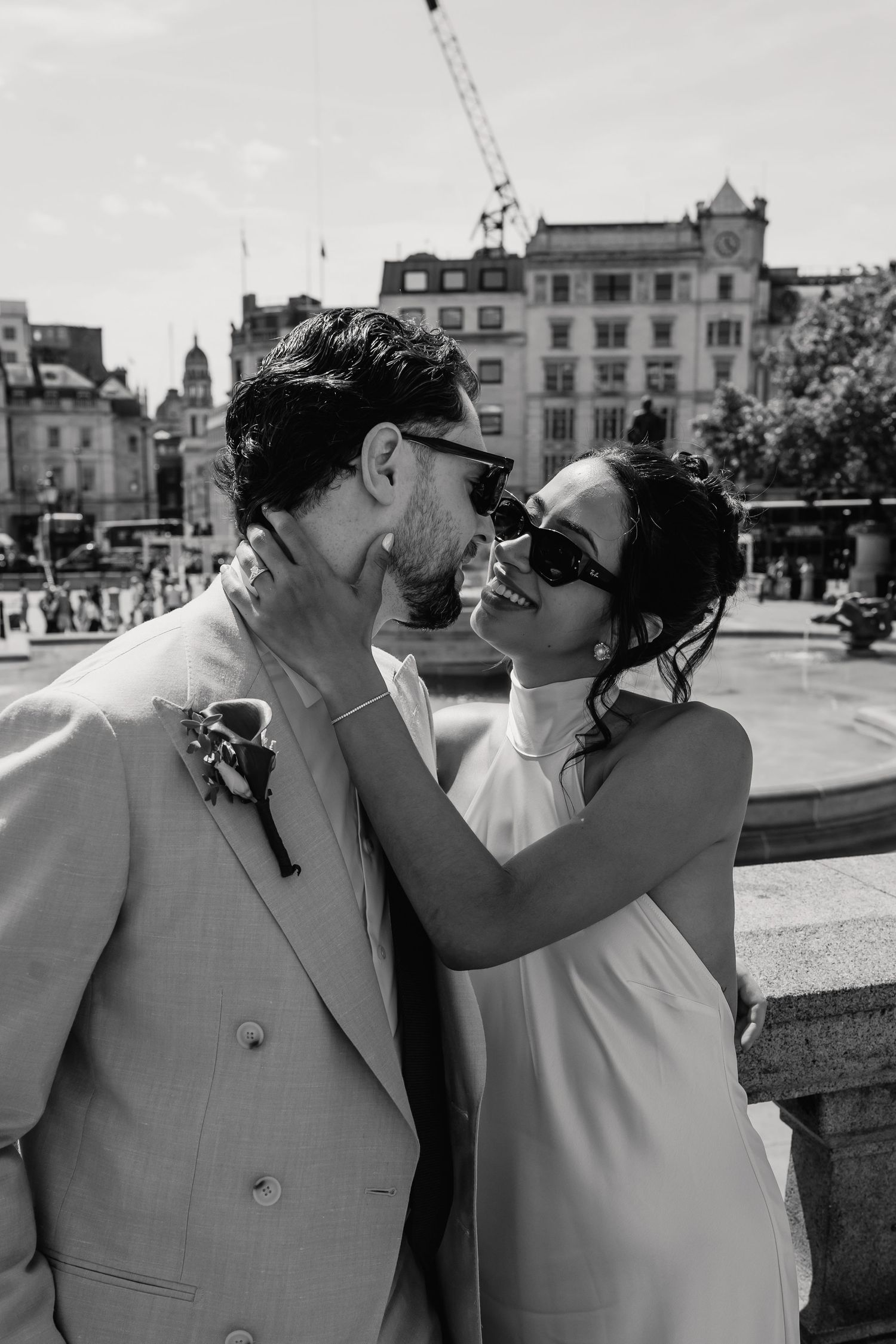 Black and white photo of bride and groom in sunglasses sharing a romantic moment by Trafalgar Square fountain.