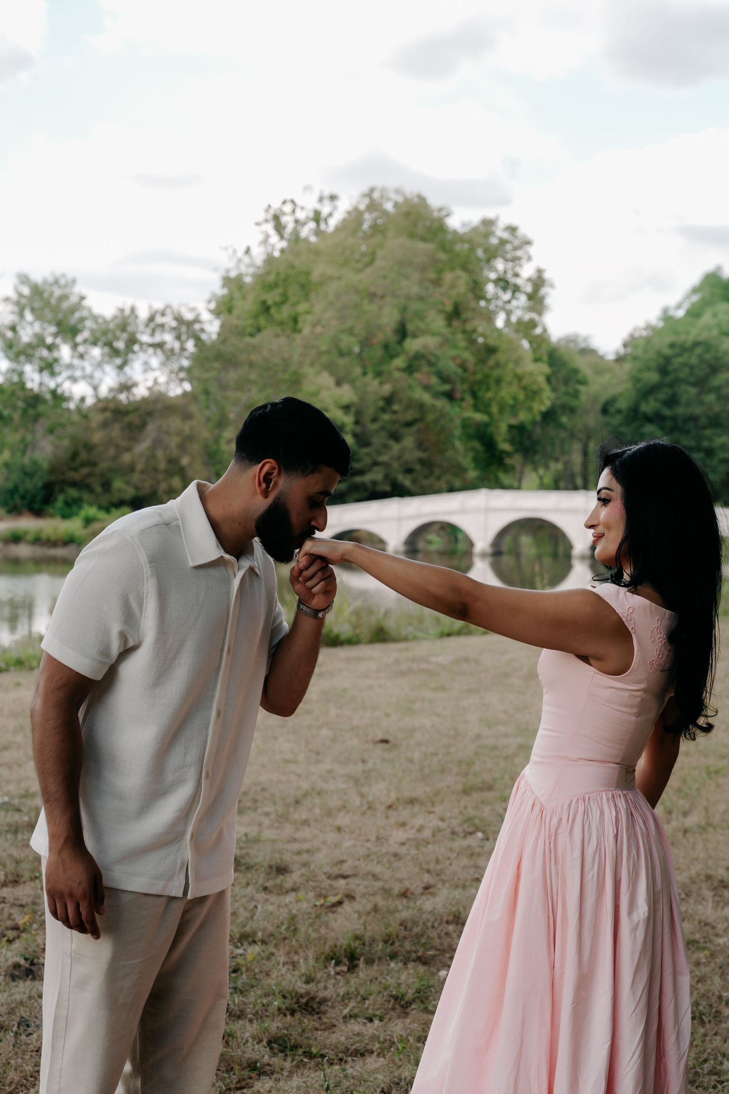 Man kisses woman's hand as she smiles in a pink dress, standing in a park with a stone bridge behind them.