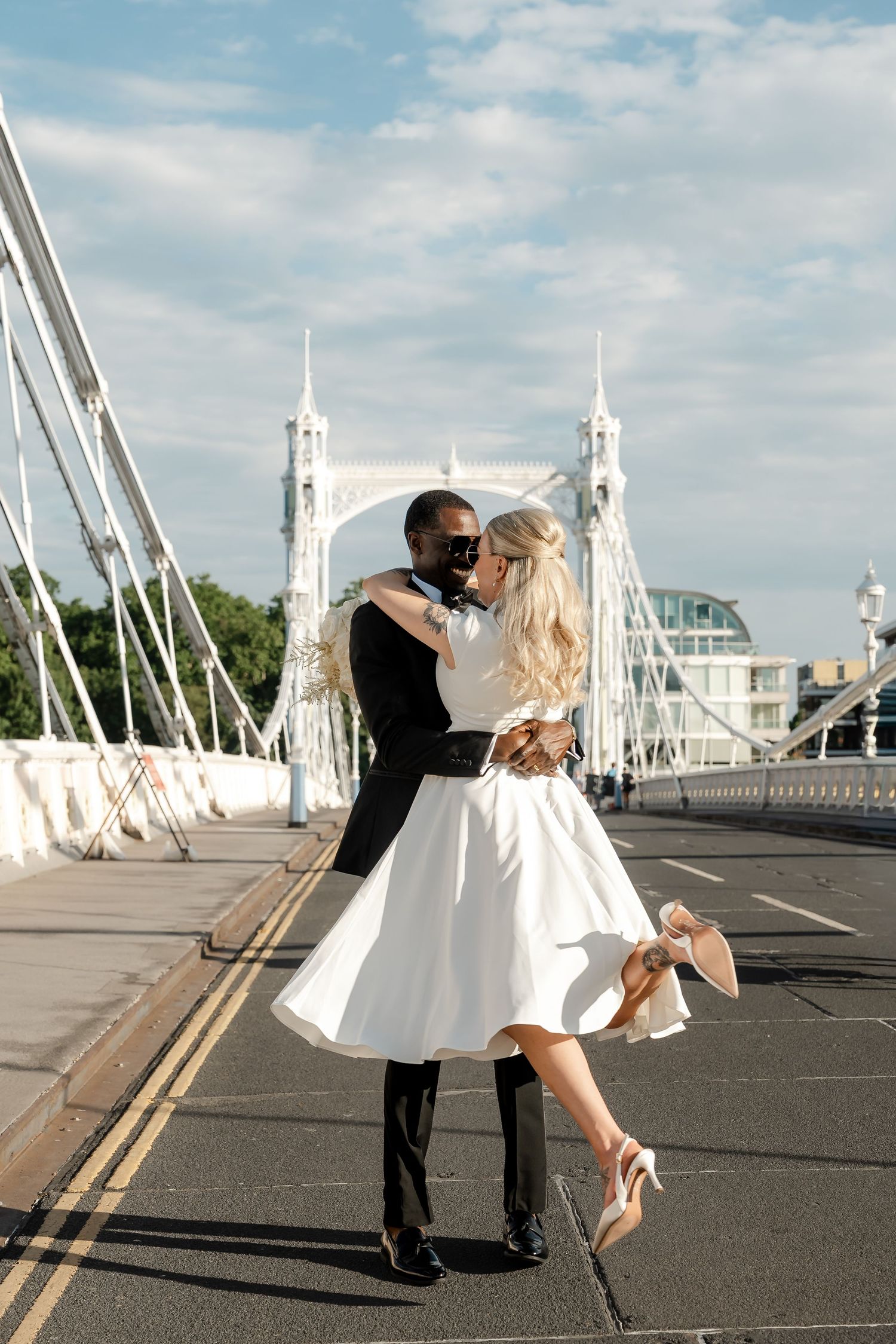Interracial couple sharing a romantic kiss on Albert Bridge London, bride in white midi dress with groom in black suit.