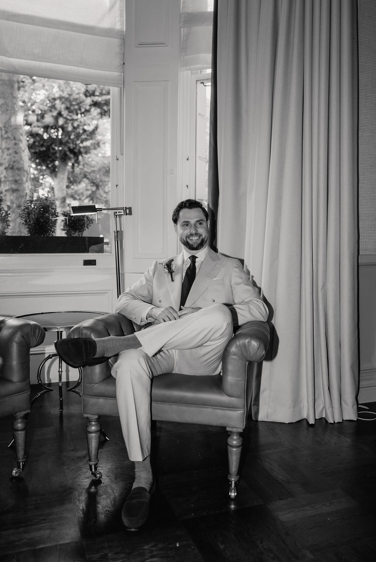 Stylish groom in light suit sitting relaxed in leather chair smiling on wedding day.