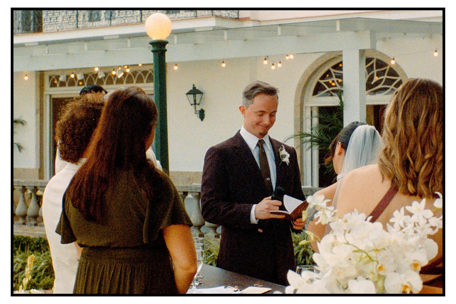 Wedding officiant reading vows at outdoor ceremony with string lights and elegant venue backdrop.