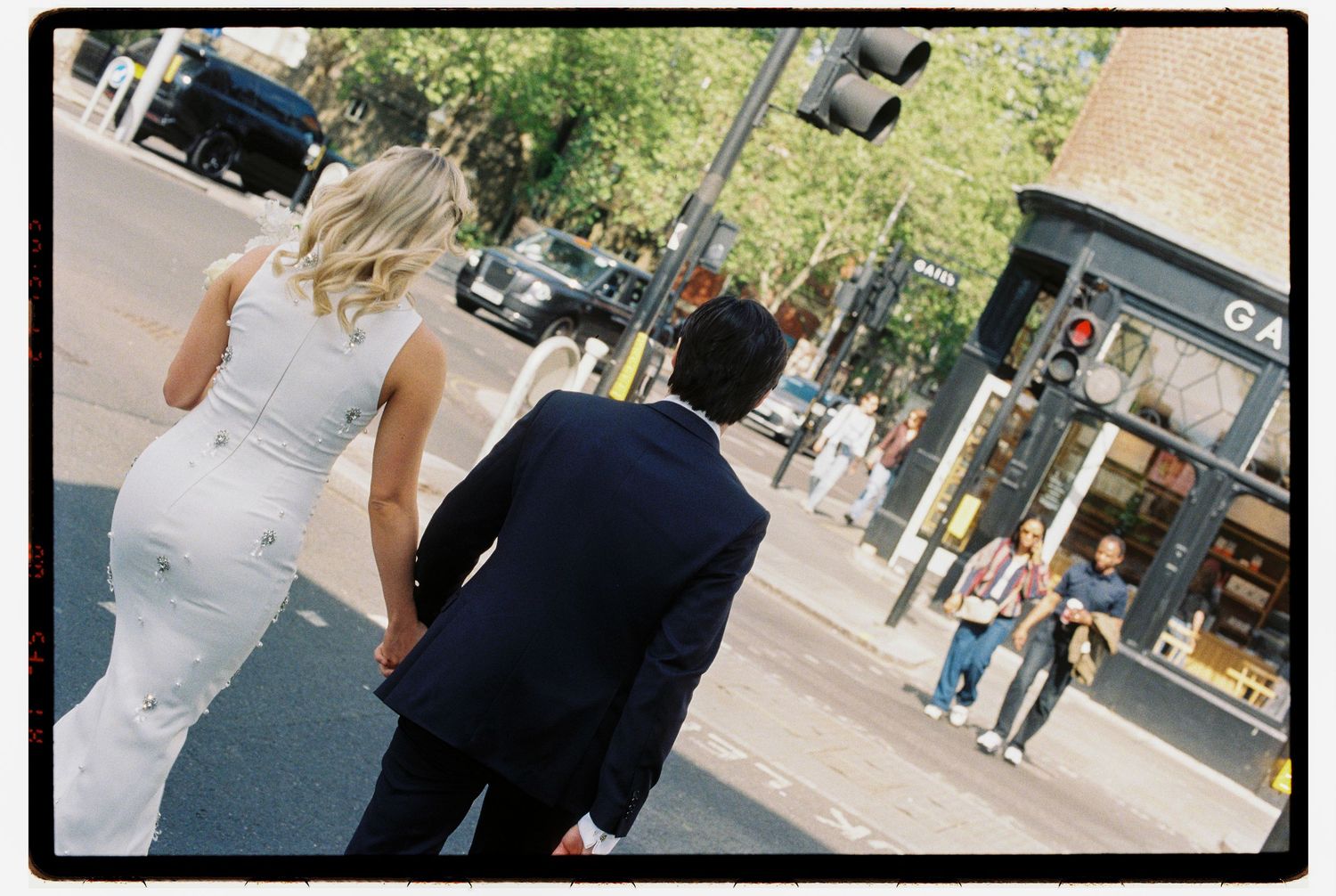 Bride in white dress and groom in navy suit walking hand in hand across a sunny city street.