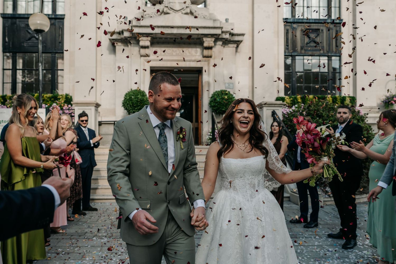 Joyful newlyweds walk through confetti shower outside elegant venue surrounded by wedding guests.