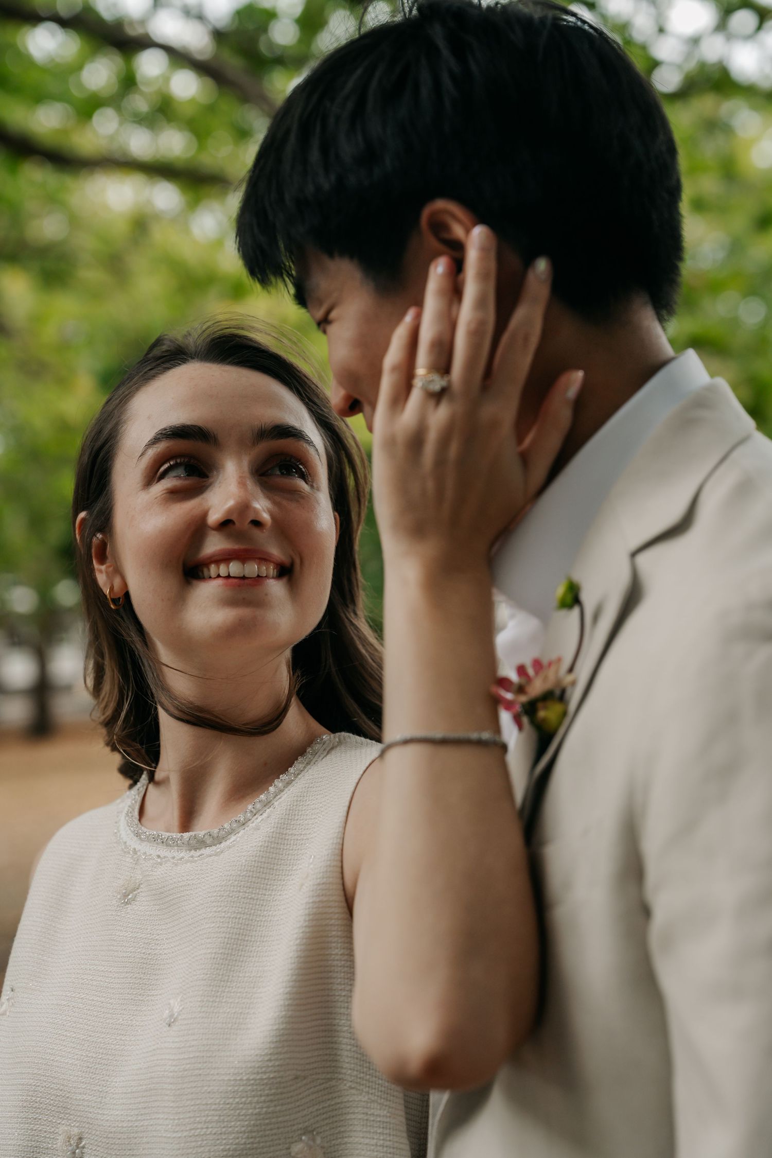 Bride smiling tenderly while gently touching grooms face outdoors under green trees.
