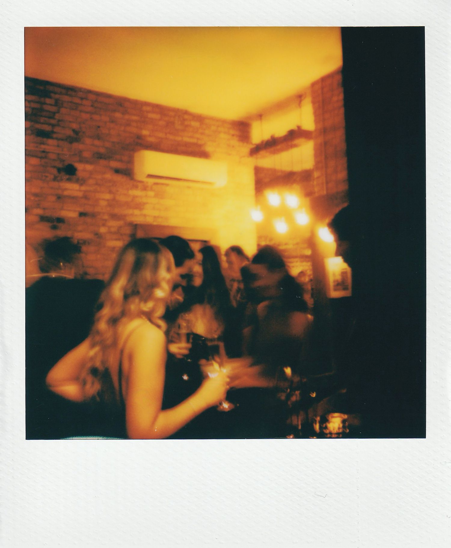 Blurry Polaroid photo of women holding wine glasses at a dimly lit bar with warm golden lighting and brick walls.