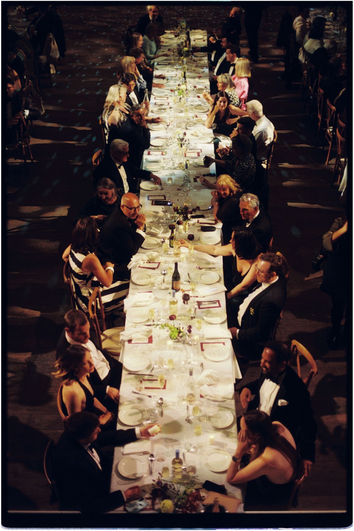 Aerial view of elegantly dressed guests dining at a long banquet table set with white linens and candles.