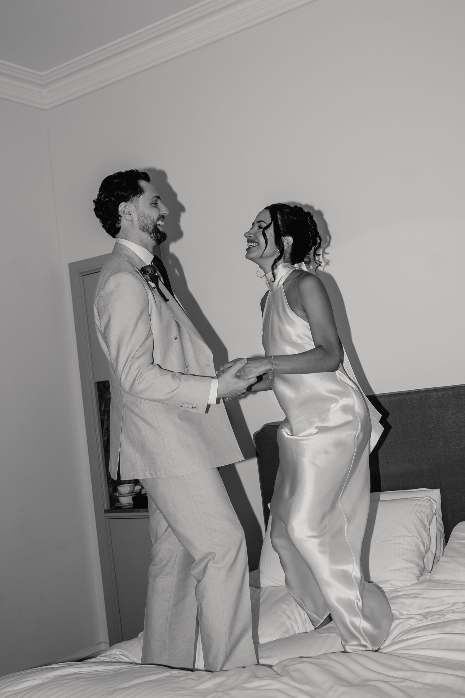 Black and white photo of laughing newlyweds jumping on a hotel bed, bride in silk dress and groom in suit.
