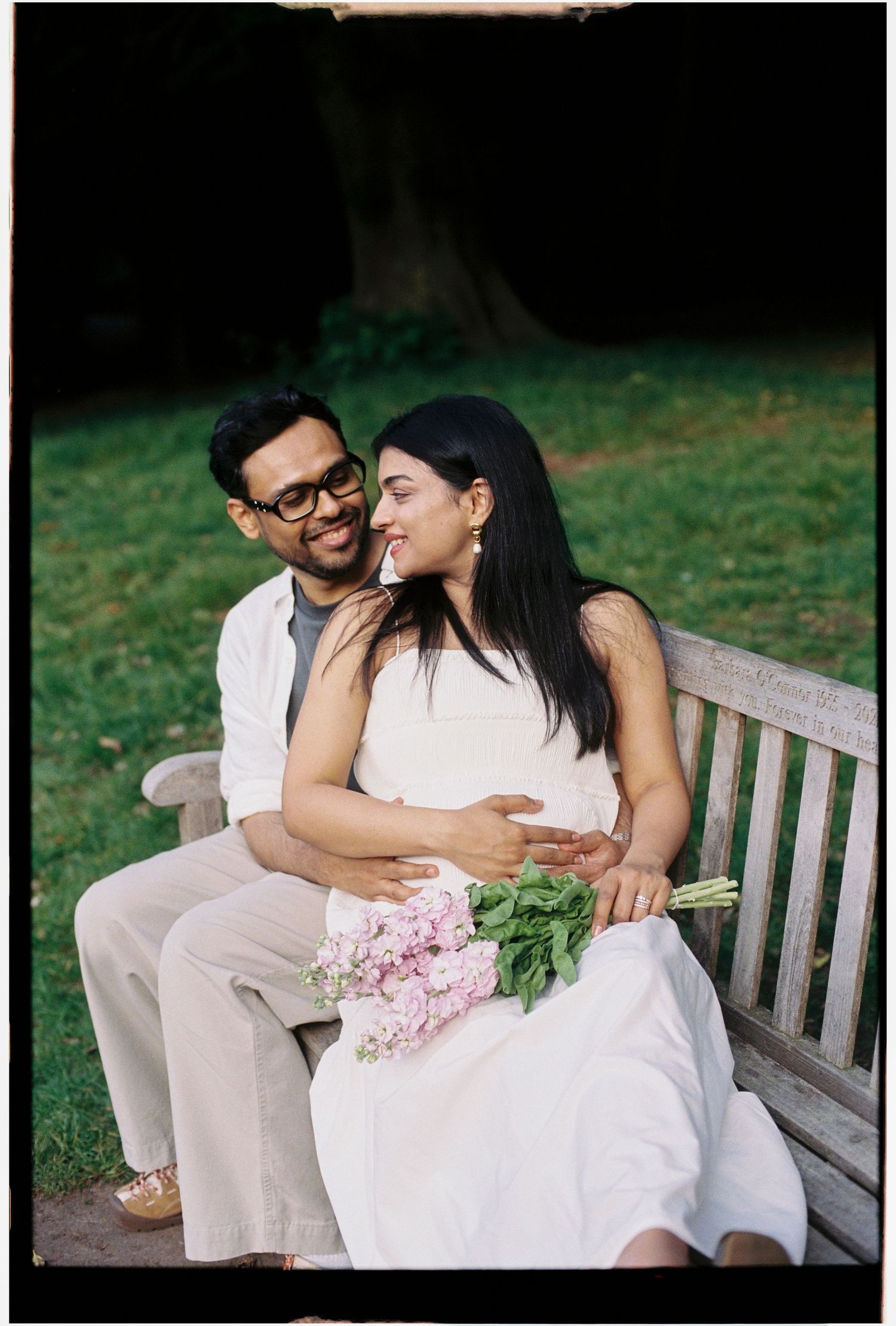 Couple in love sit on a garden bench, bride holding pink flowers in a park setting.
