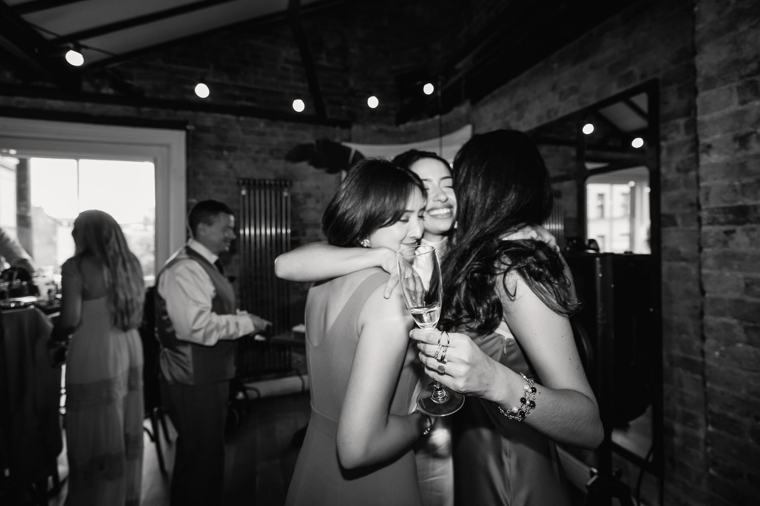 Two female wedding guests embrace warmly while holding champagne glasses at the reception.