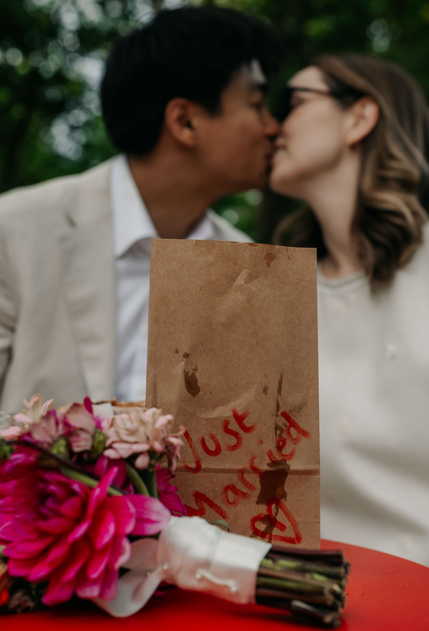Just married couple kissing behind brown paper bag sign with vibrant pink floral bouquet.