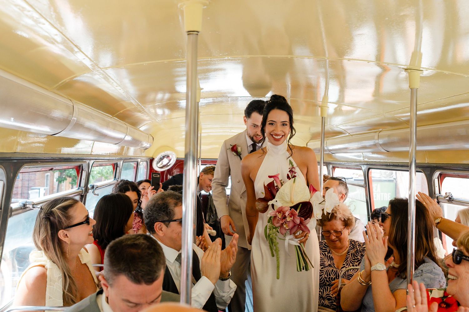 Bride in white halter dress and groom standing in aisle of vintage bus as wedding guests celebrate around them.