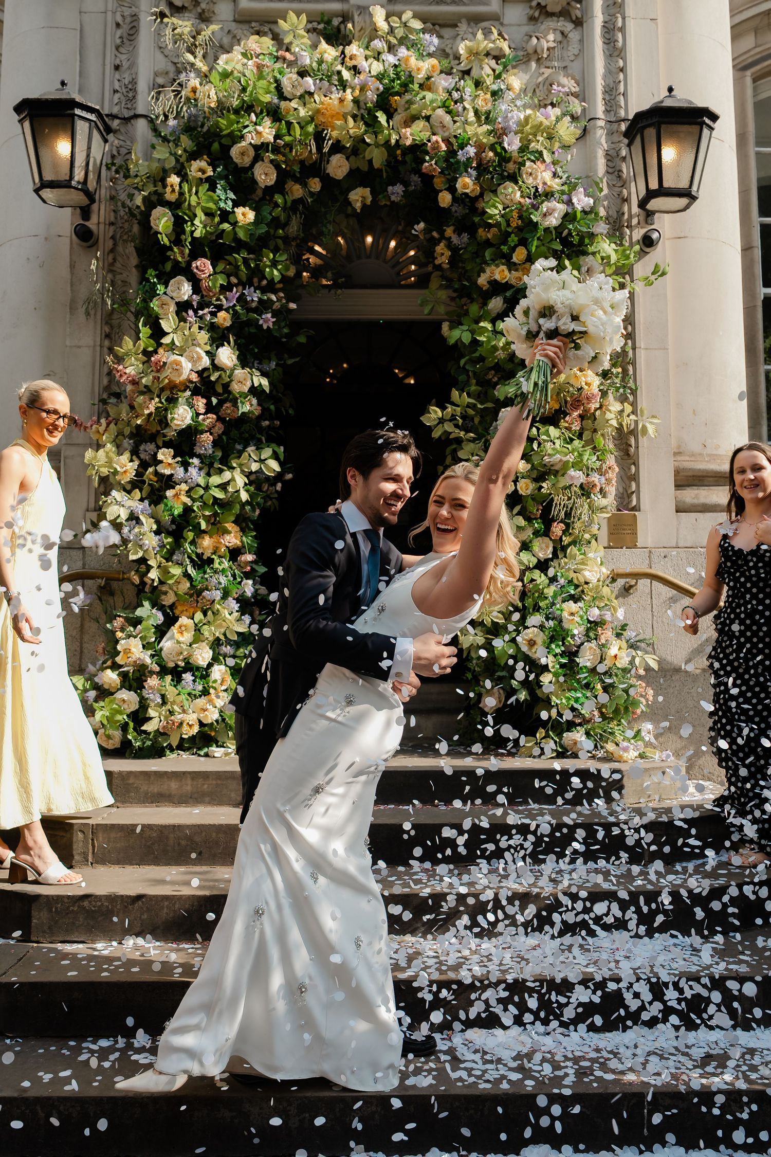 Bride and groom celebrate with confetti outside a floral-adorned wedding venue doorway.