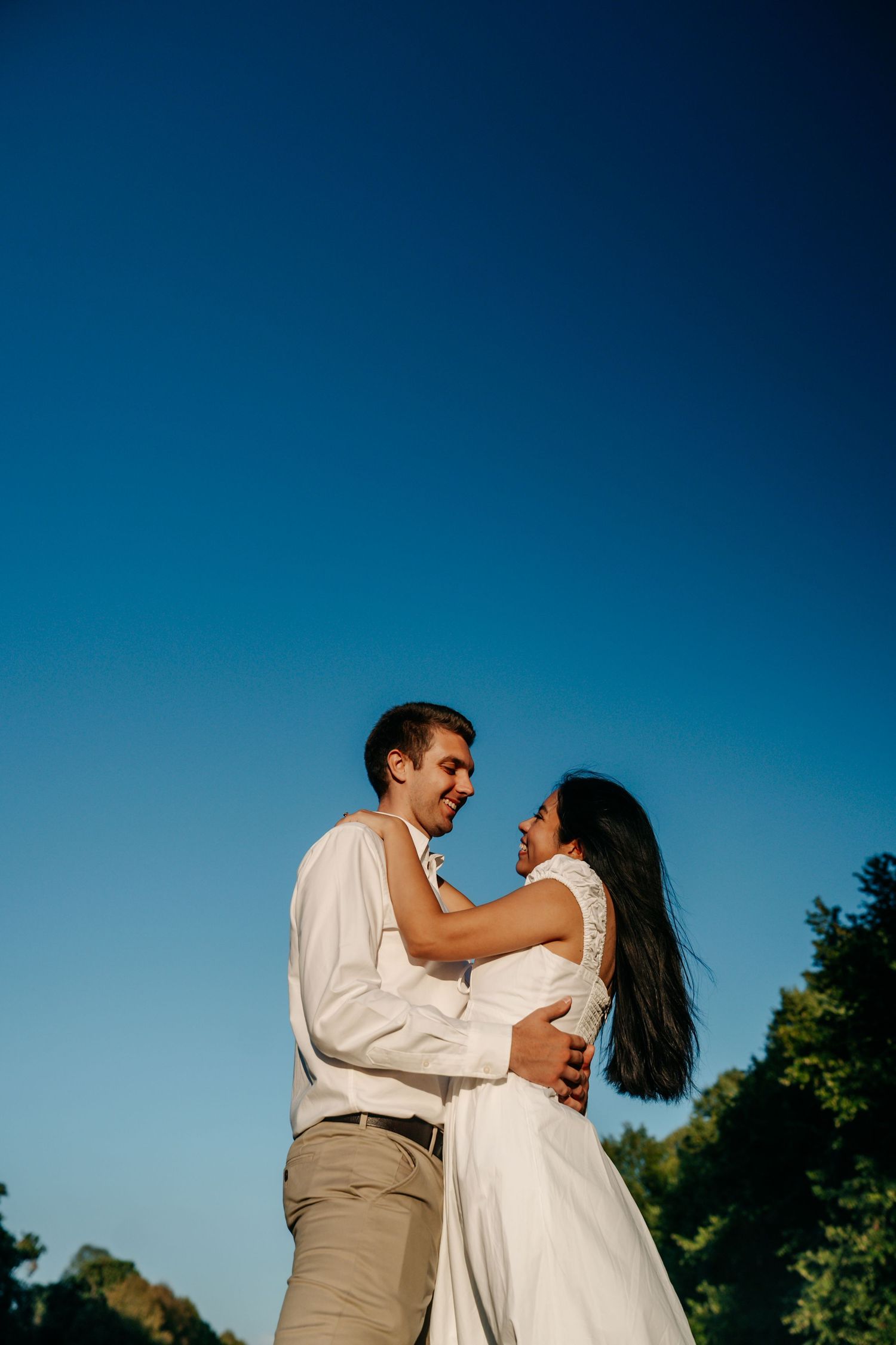 Happy couple embracing and smiling at each other against a vivid blue sky, woman in white dress, man in white shirt.