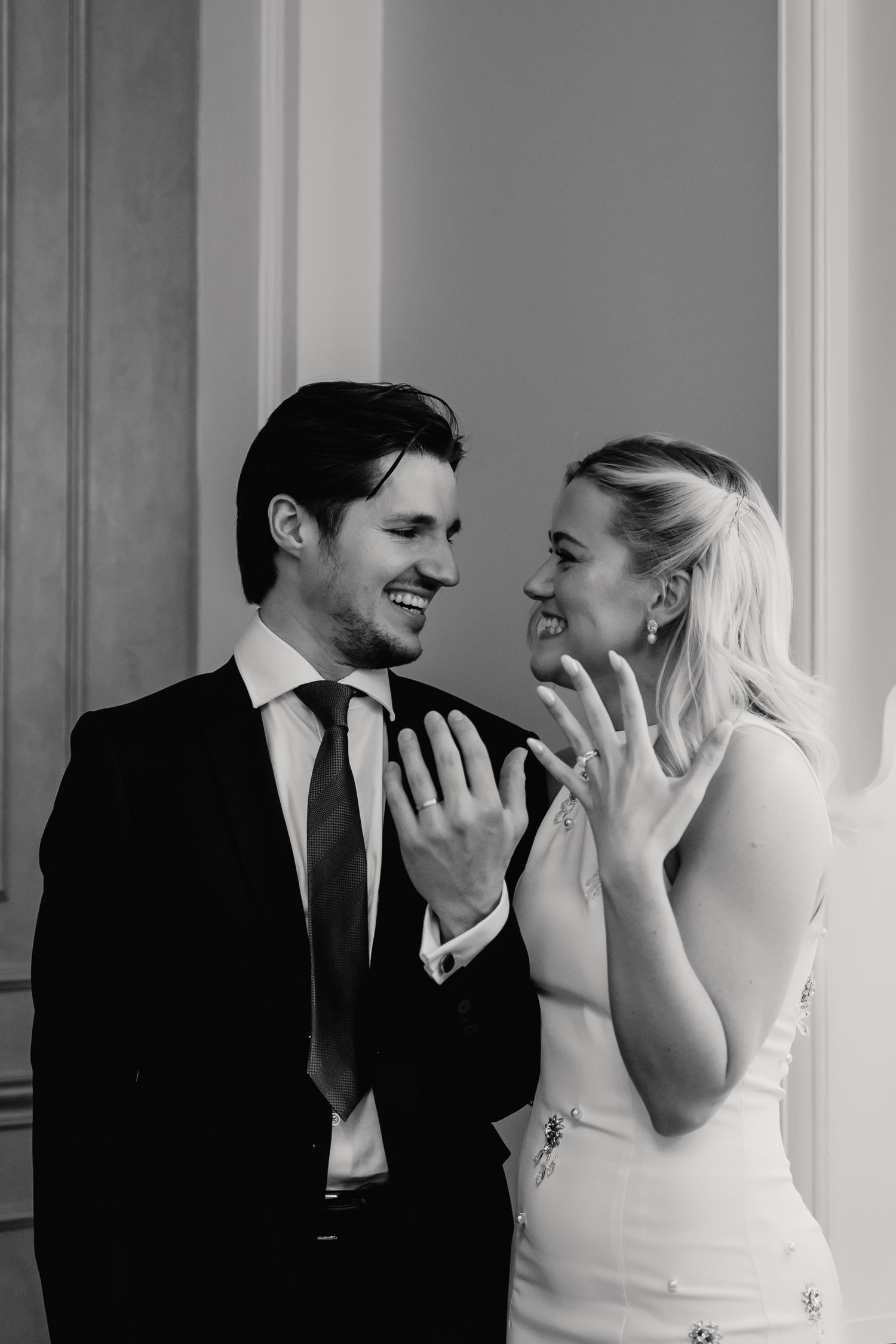 Black and white photo of smiling newlyweds proudly displaying their wedding rings to the camera.