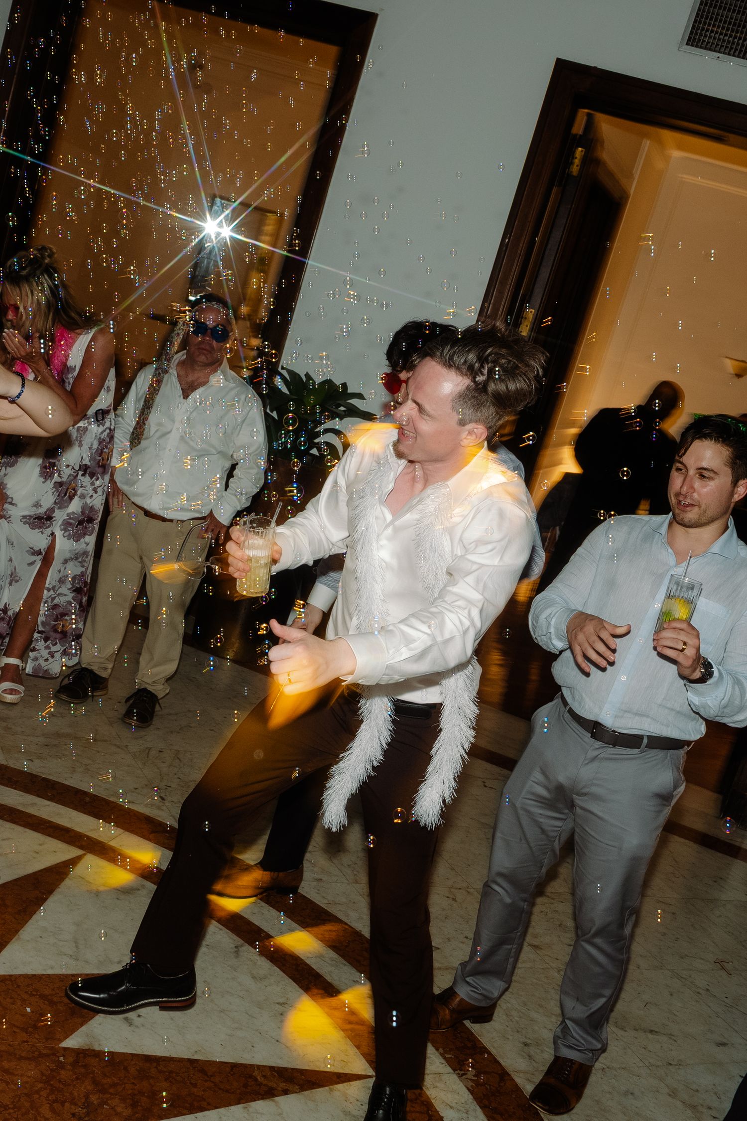 Man in white shirt with feather boa dances energetically at glittering wedding reception party.