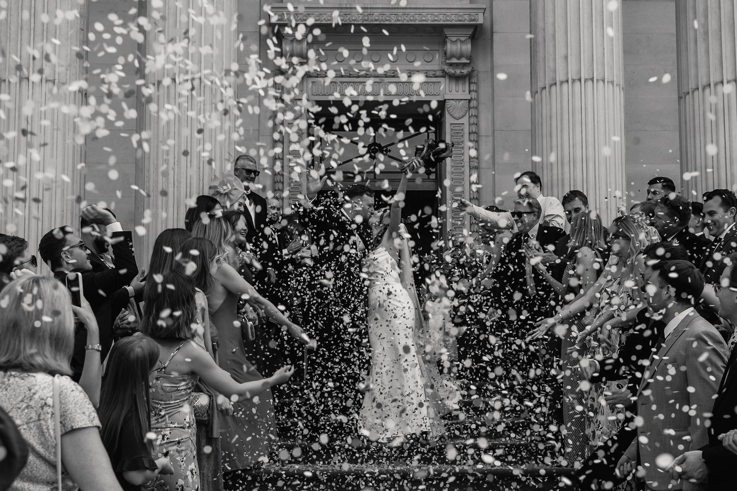 Black and white photo of bride and groom walking through shower of confetti thrown by wedding guests.