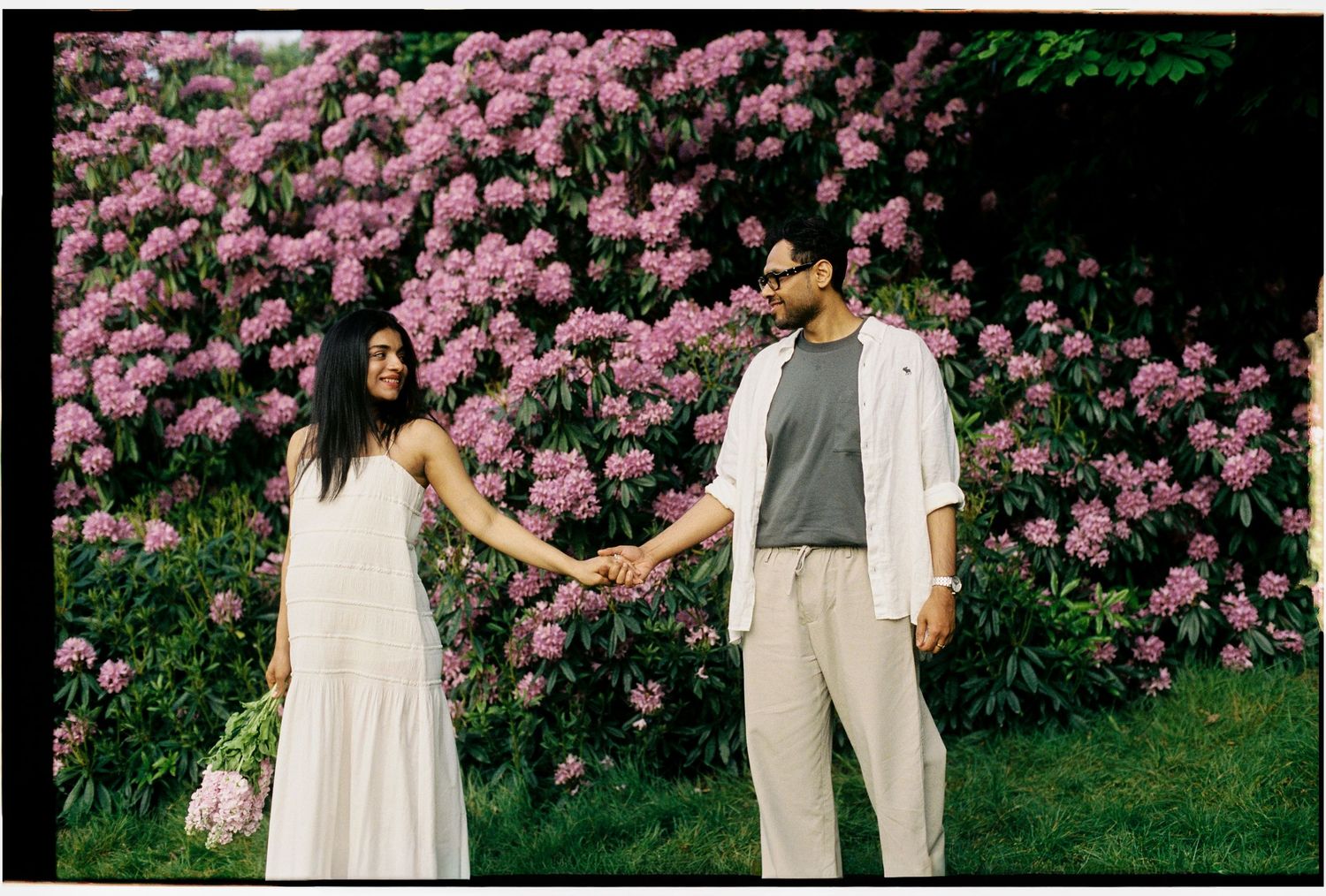Couple holding hands in front of pink rhododendron blooms, woman in white dress, man in casual white outfit.