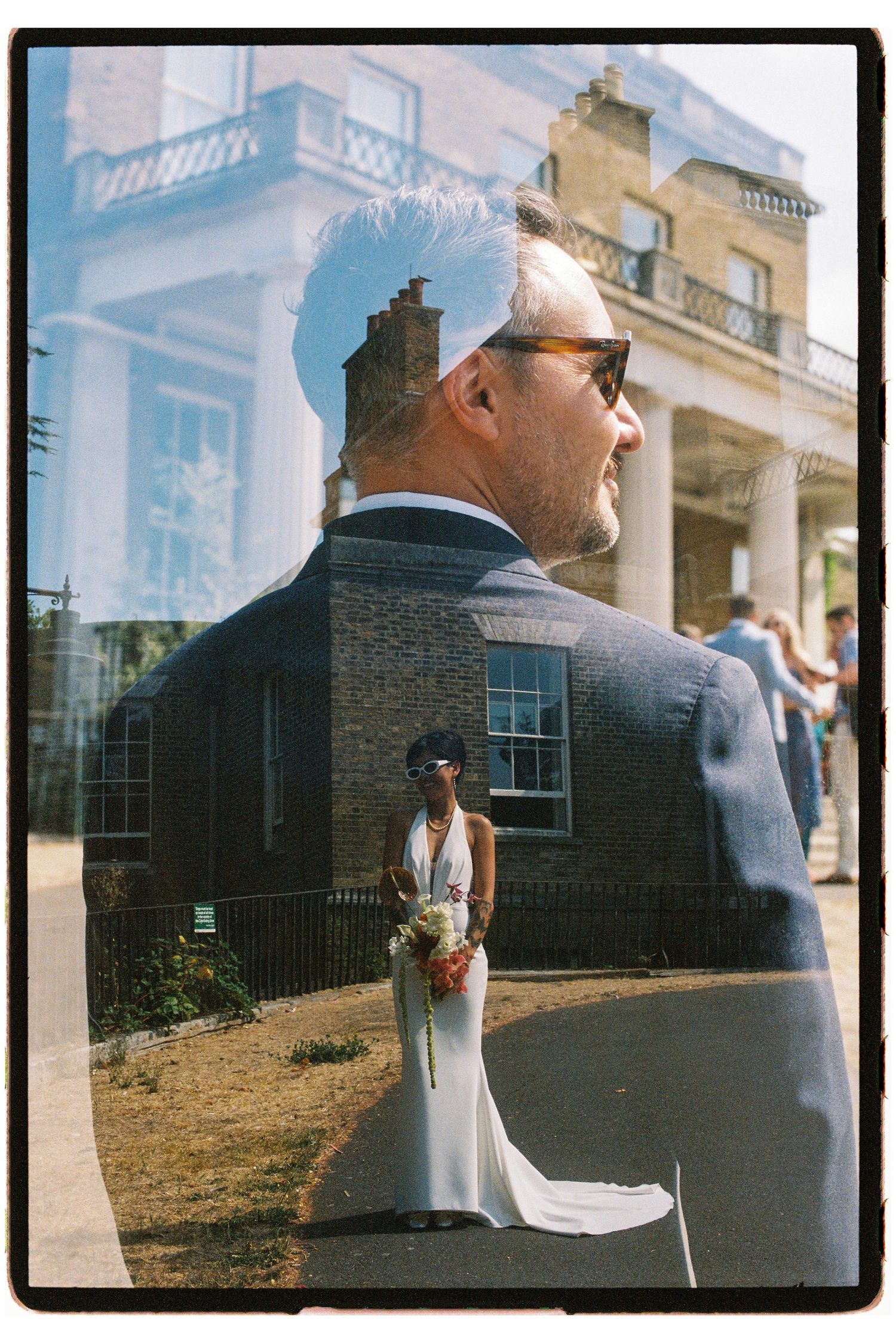 A creative double exposure film photo of a groom and bride in white holding a bouquet outside a historic building.