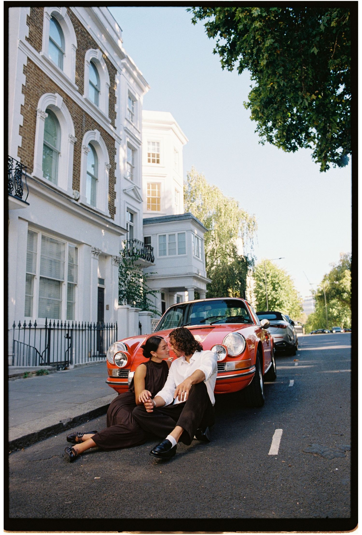 Couple kissing on a street in London beside a vintage red Porsche, surrounded by classic architecture.