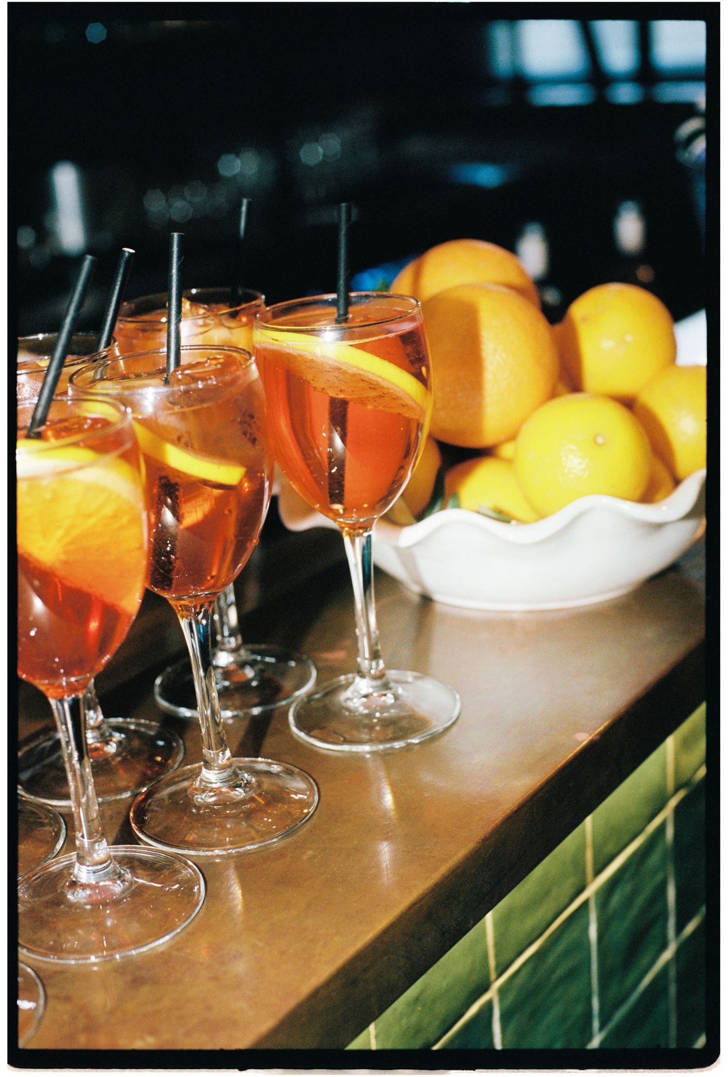 Four Aperol Spritz cocktails with orange slices and straws on a bar counter beside a bowl of fresh oranges.