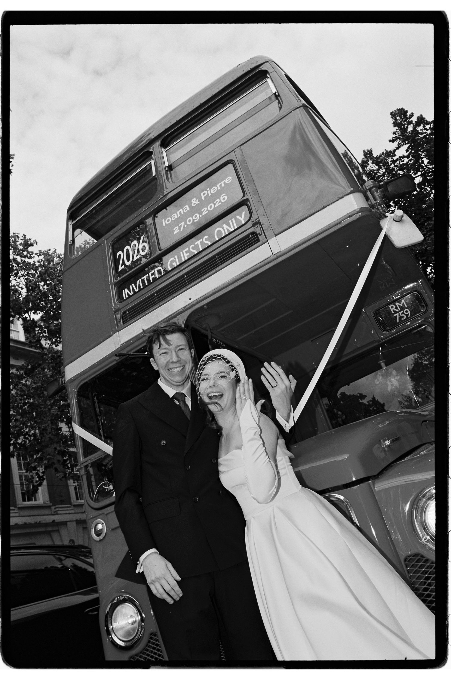 A joyful couple in wedding attire poses in front of a vintage double-decker bus in a black and white photo.