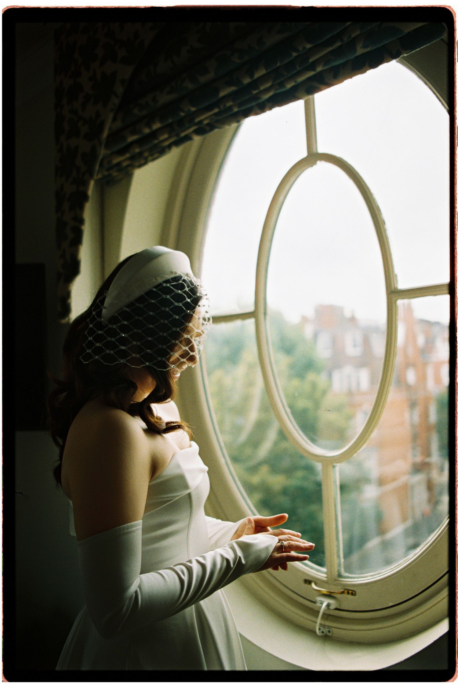 Bride in white gown and birdcage veil gazes out an oval window at a lush garden view on her wedding day.