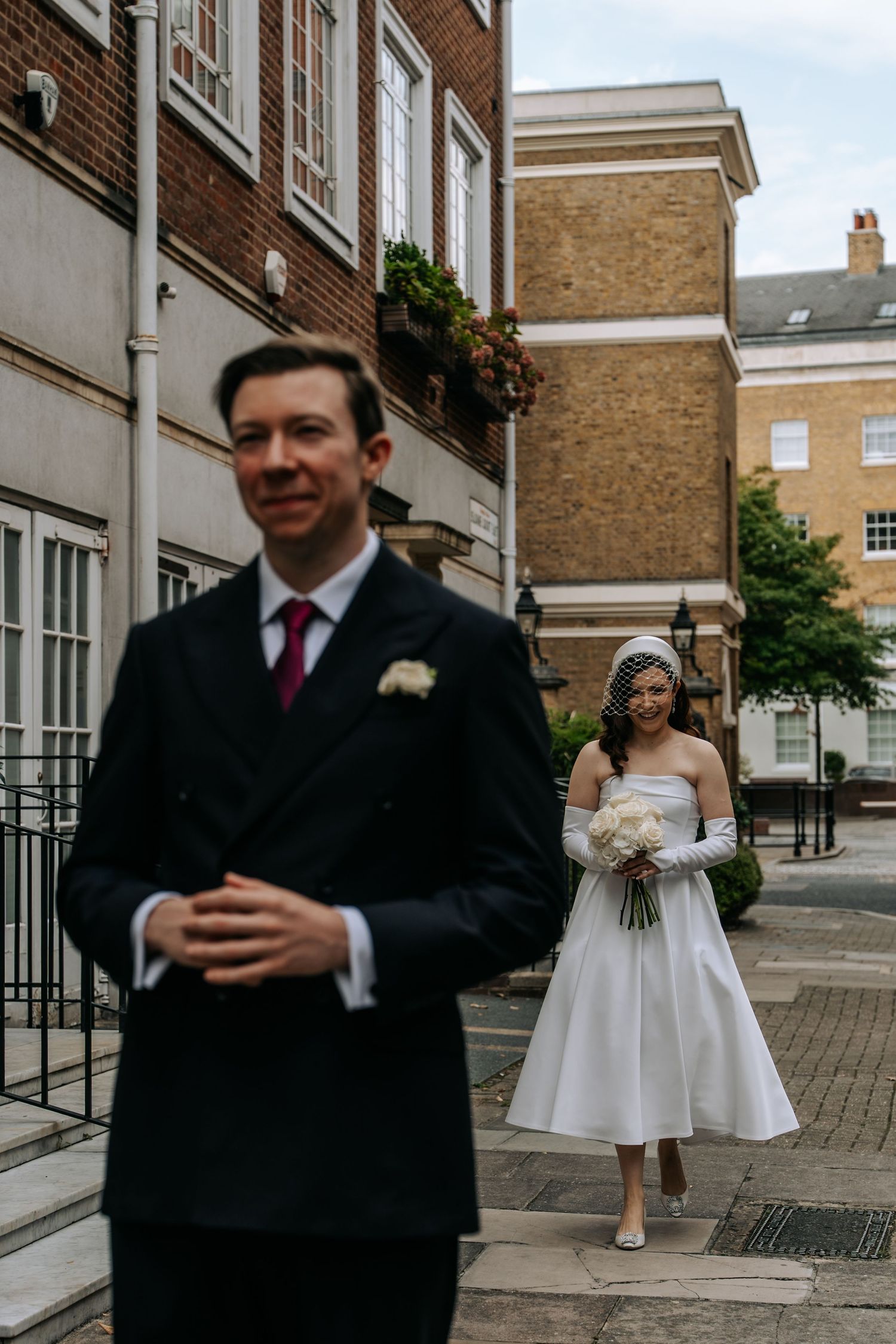 Bride in tea-length white dress and veil walks toward groom in dark suit on cobblestone street outside brick building.