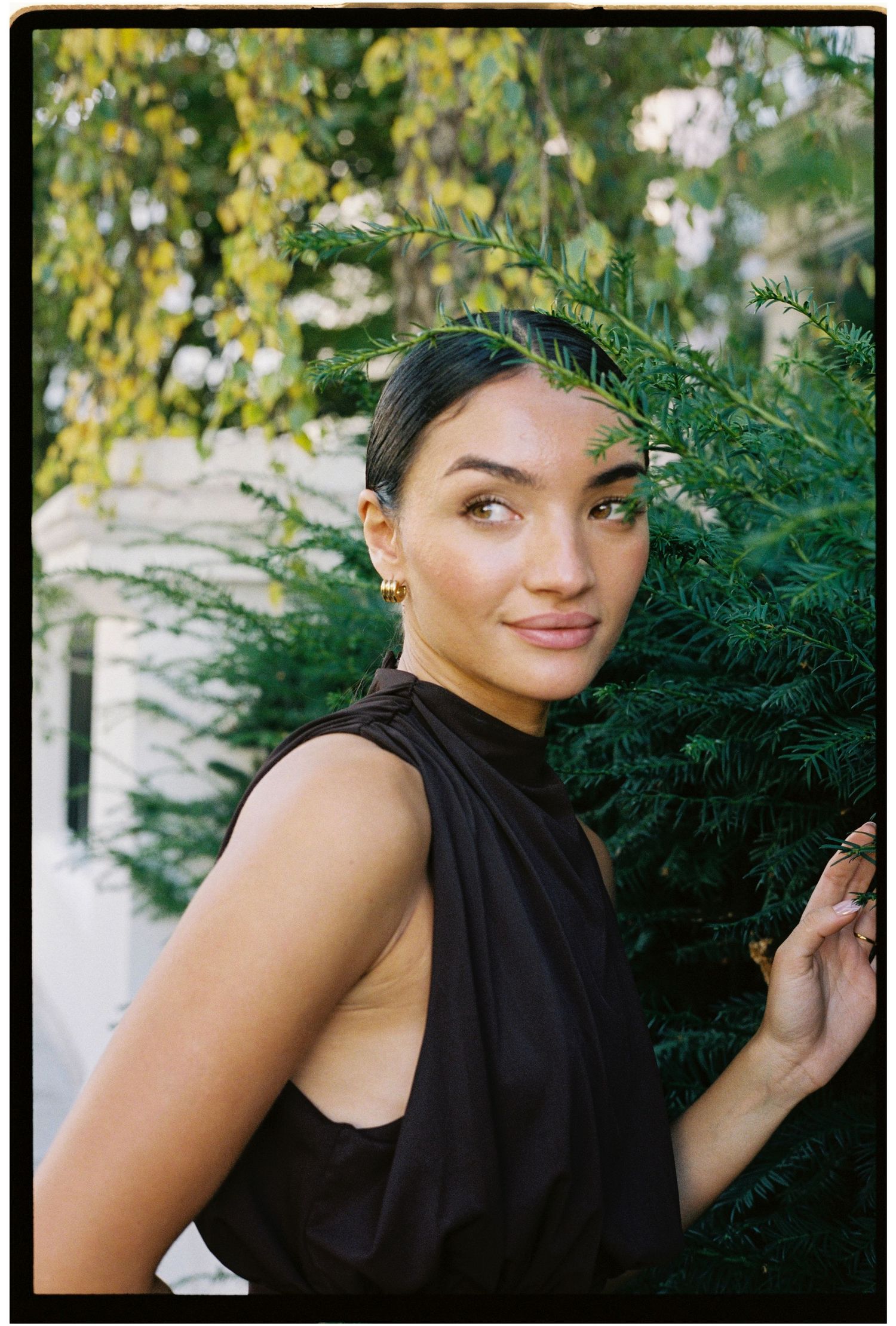 Young woman in black sleeveless top posing outdoors beside lush green evergreen trees, looking over shoulder.