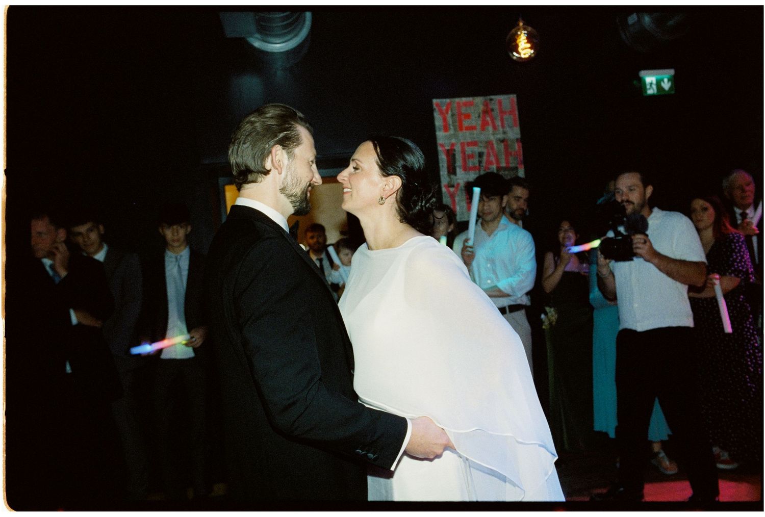 A couple shares a first dance kiss at their wedding reception while guests celebrate around them in a dimly lit venue.