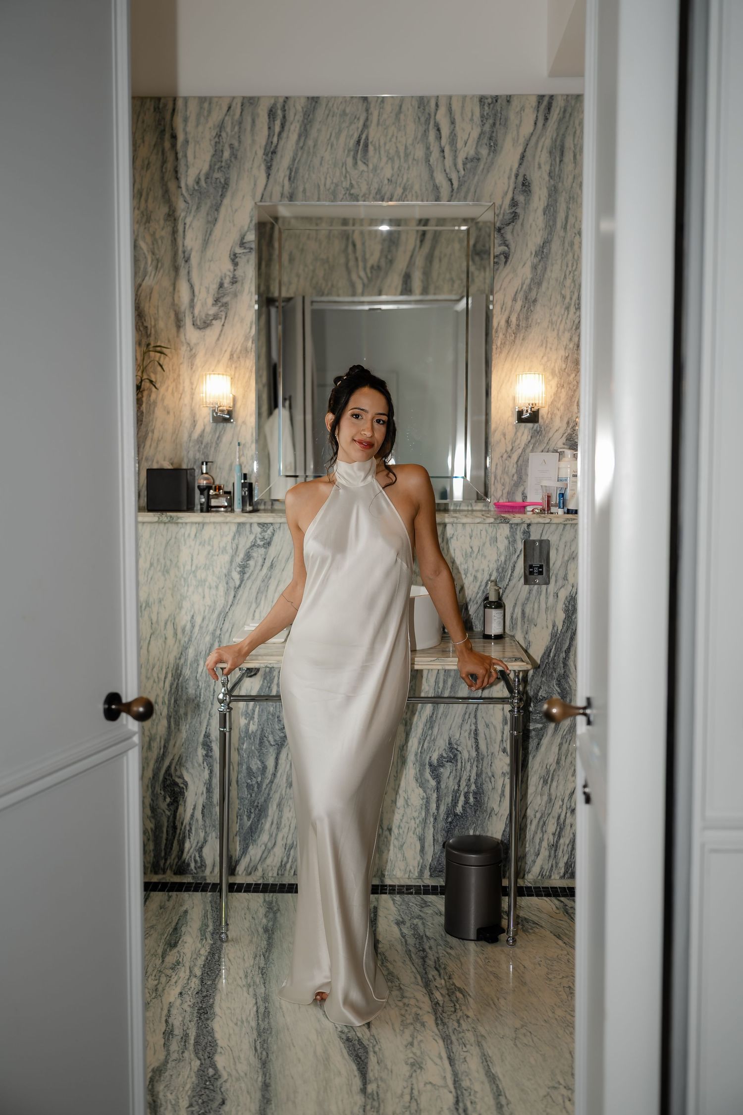 Elegant bride in a white silk halter gown posing in a stunning marble bathroom doorway on her wedding day.