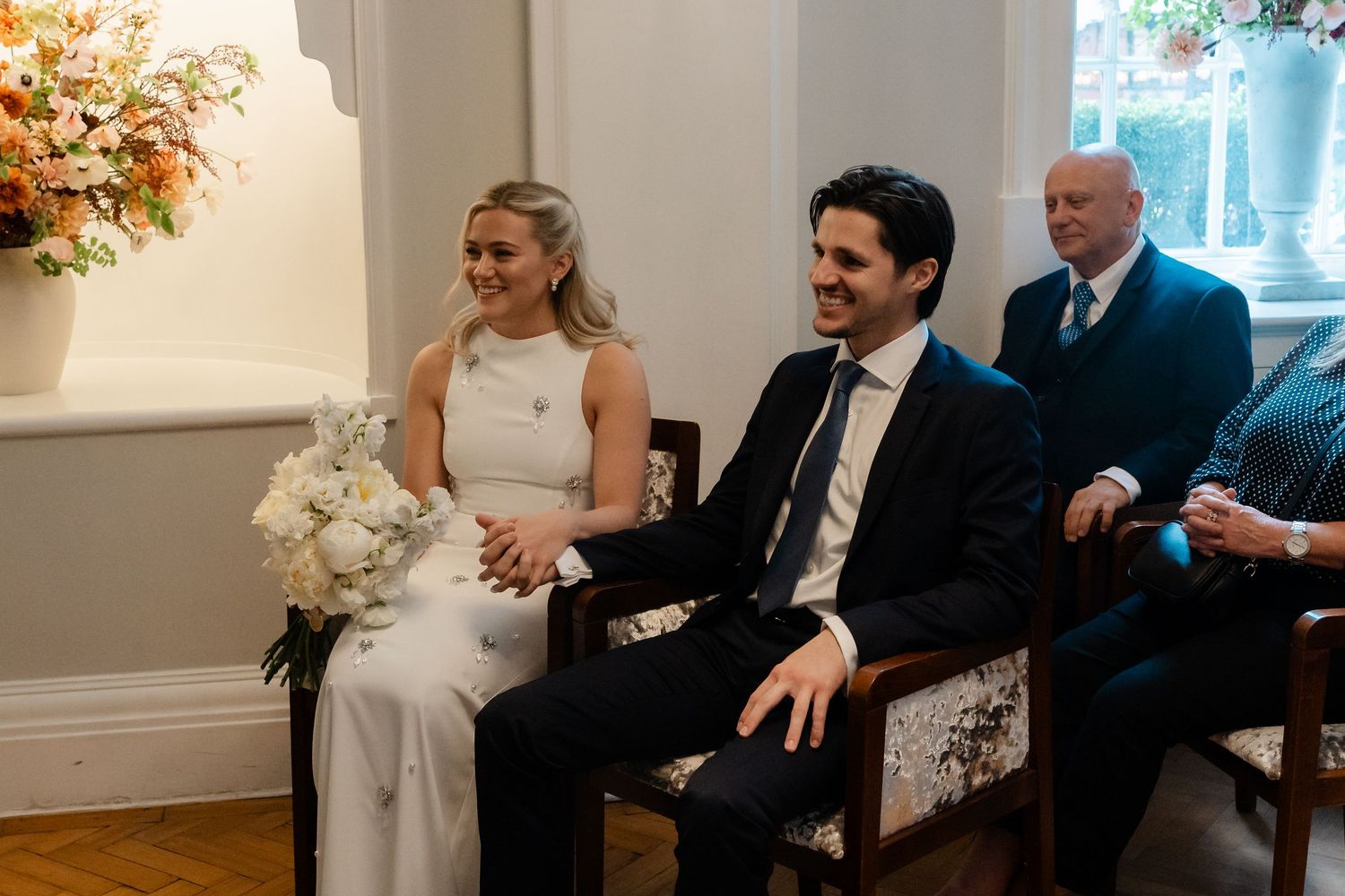 Bride holding white bouquet and groom sit together smiling during their intimate wedding ceremony.