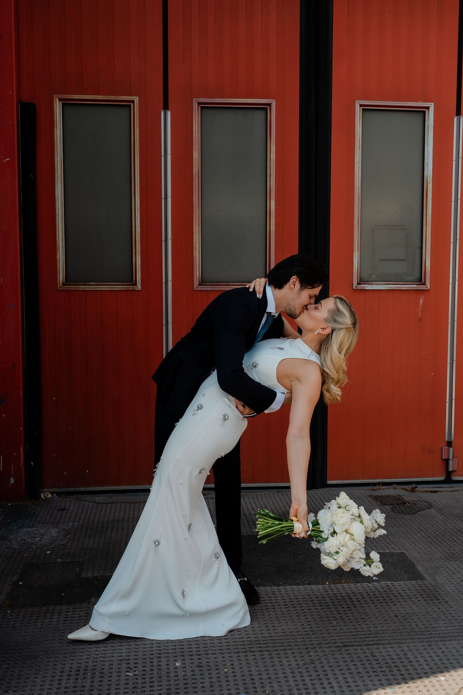 Groom dips bride in a romantic kiss against bold red doors of Chelsea Fire Station.